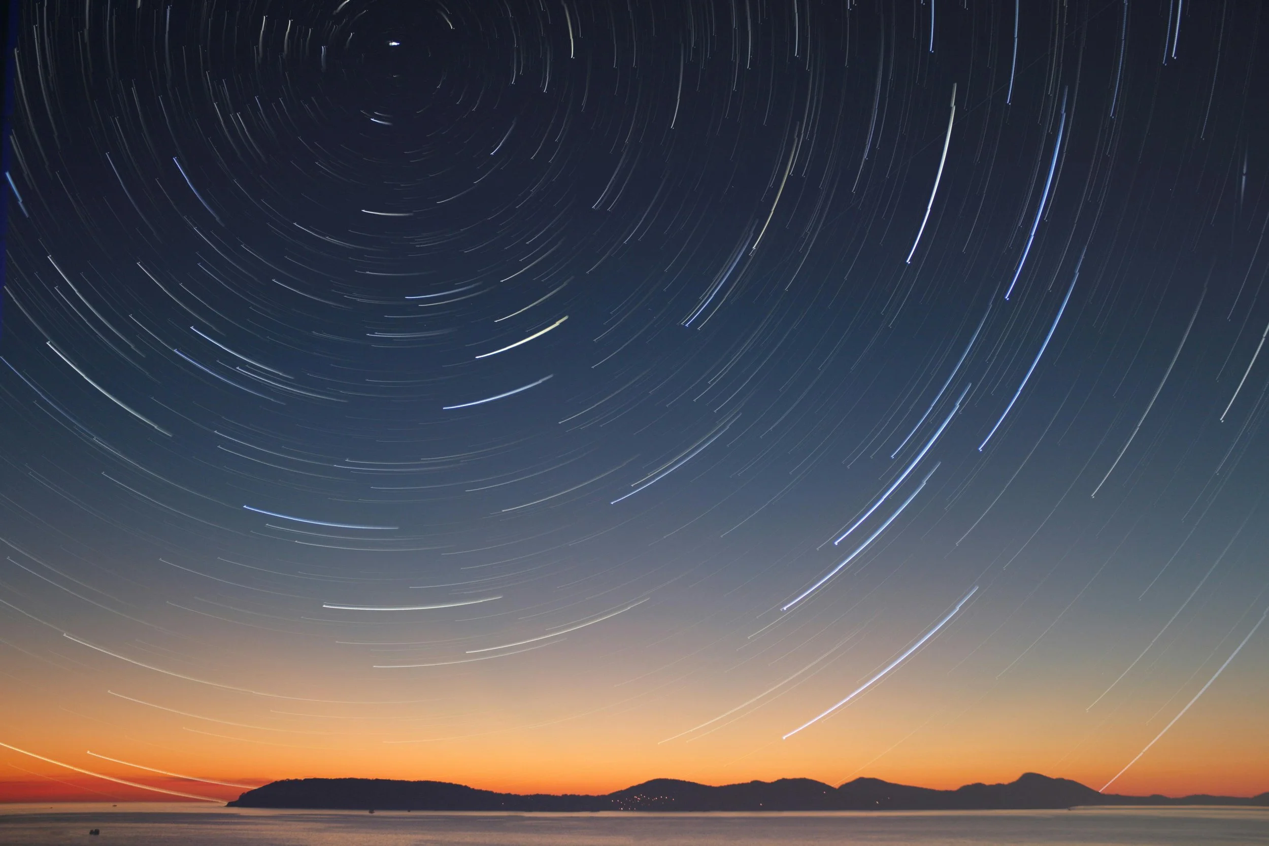 Long-exposure photograph of star trails in the night sky over a body of water with a sunset glow near the horizon.