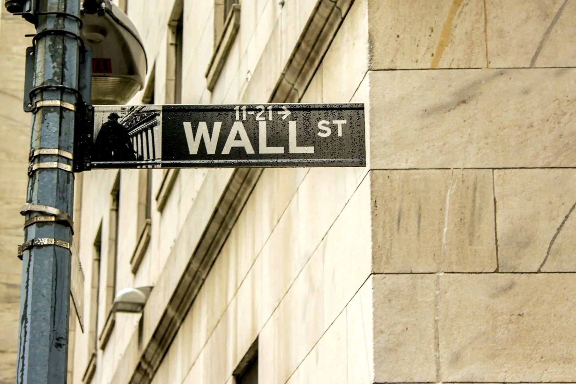 Street sign for Wall Street in New York City, showing the numbers 11-21, attached to a pole in front of a beige building with multiple windows.
