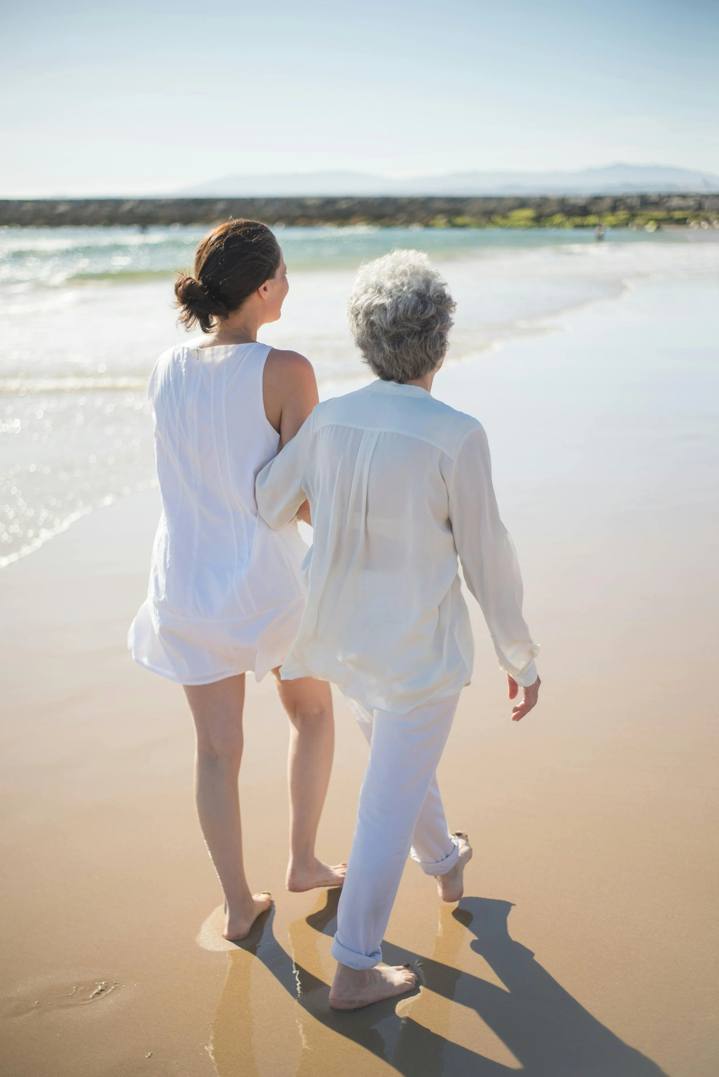Two women walking along a sandy beach near the water, on a sunny day.