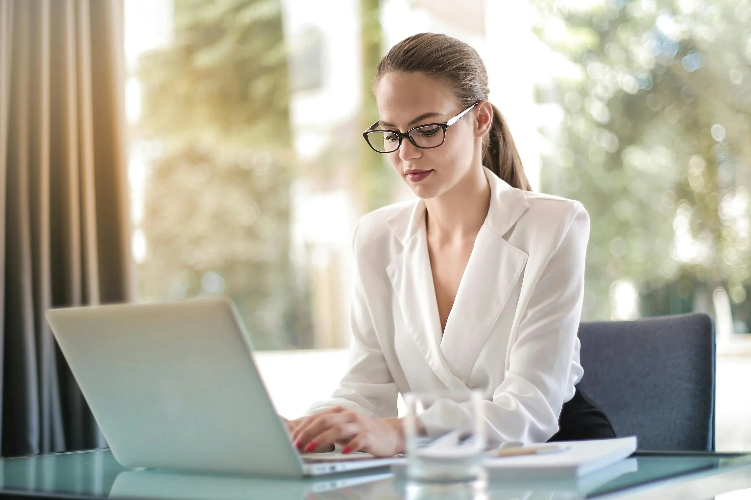 Woman wearing a white blouse and glasses with her hair tied up in a ponytail working on a laptop at her desk