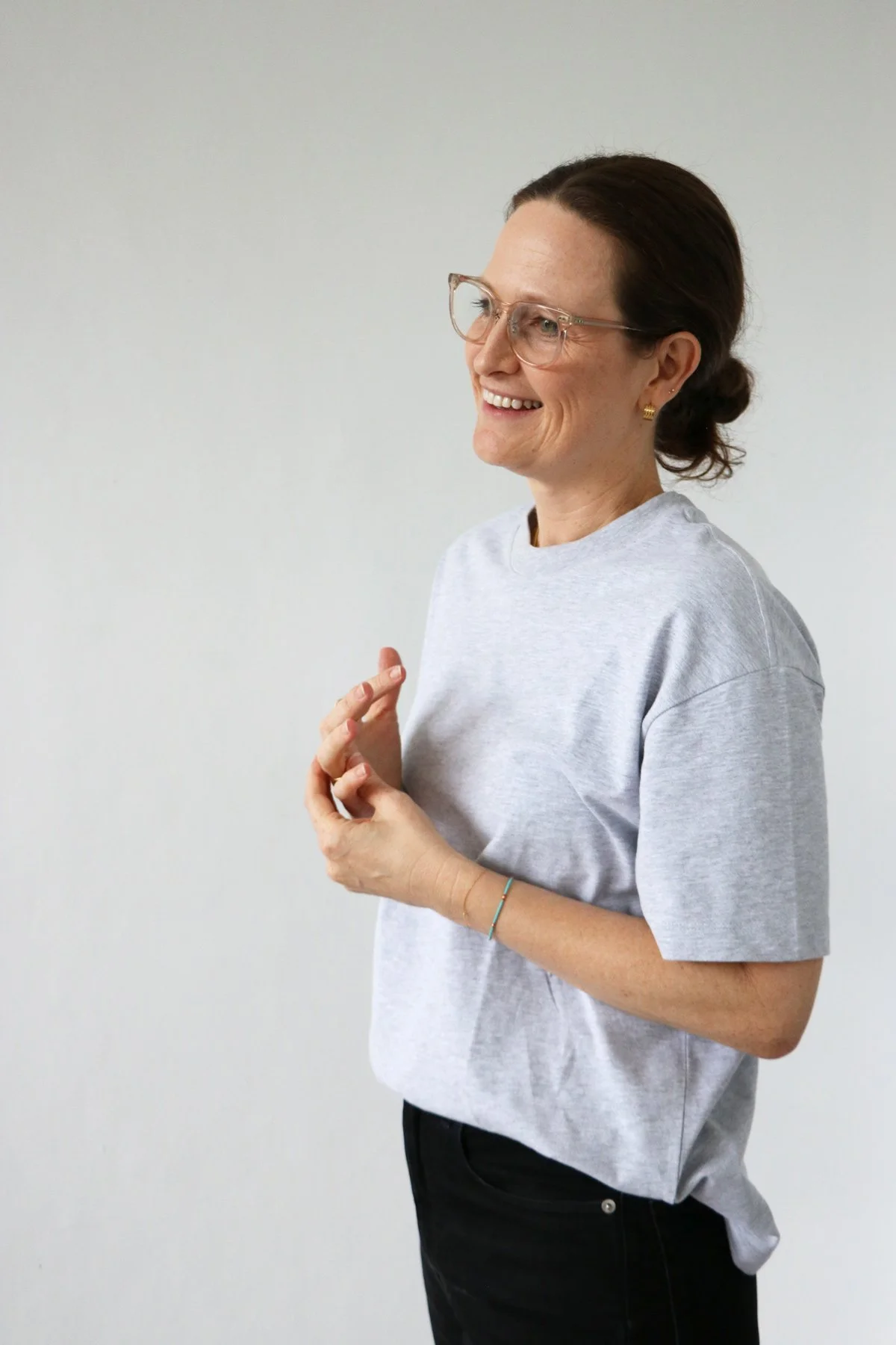 A woman with glasses and earrings smiling, wearing a light gray T-shirt, standing against a plain light-colored background.