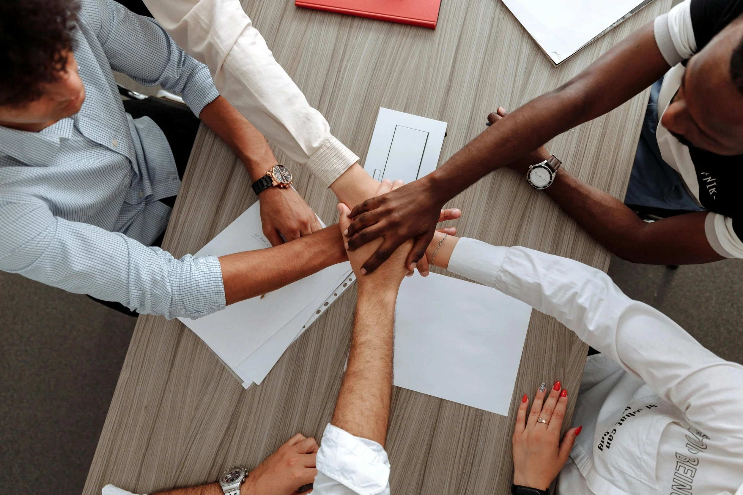 Multiple people placing their hands together in a teamwork gesture across a wooden table with papers and documents.