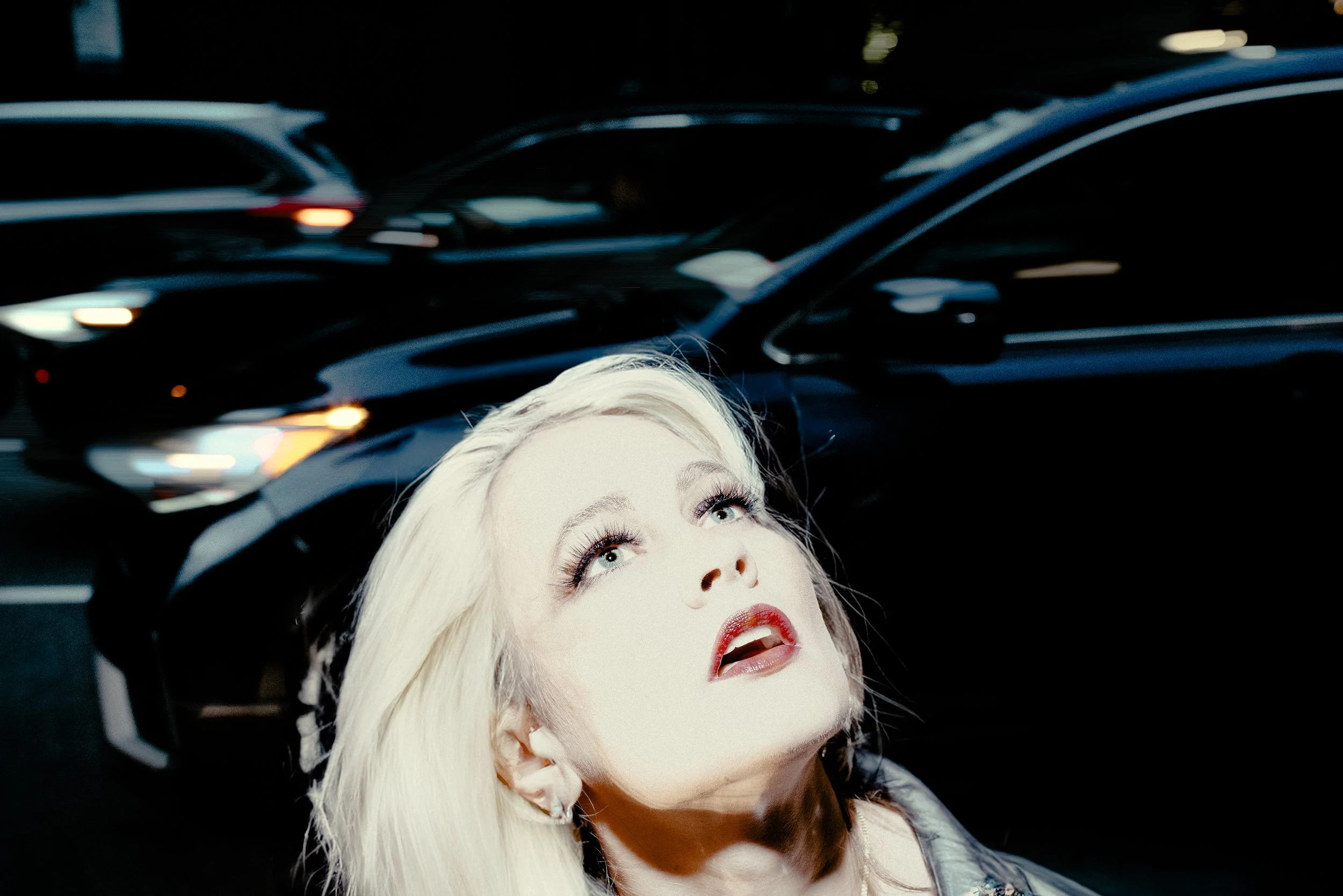A woman with platinum blonde hair and makeup, looking upwards with a surprised or curious expression, with black cars and blurred lights in the background.