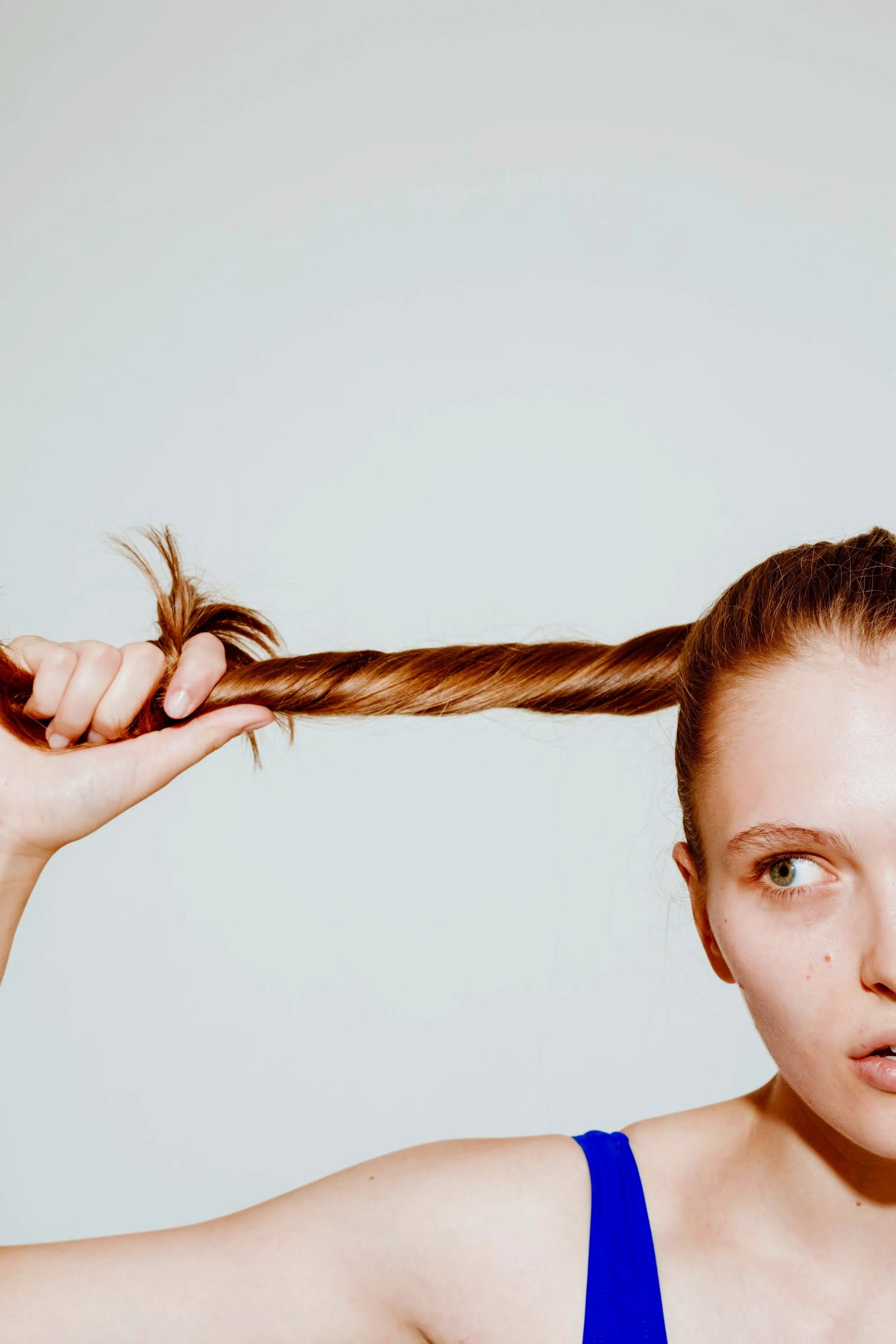 A woman with reddish-brown hair is holding her long hair out straight to the side, wearing a blue top, with a neutral expression and a light background.