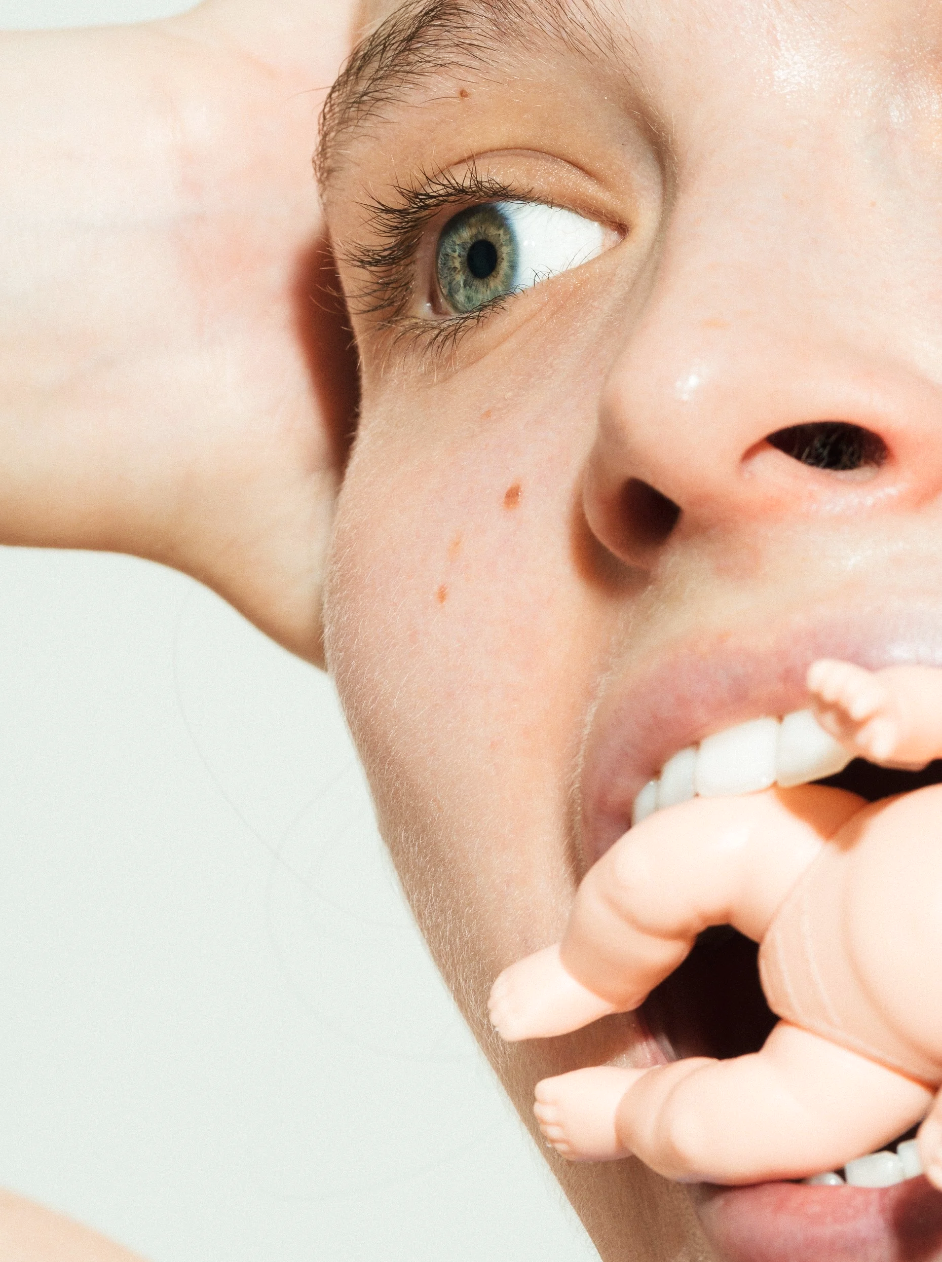 Close-up of a woman with blue eyes, pink lips, and a light skin tone, holding a pale pink silicone mannequin hand near her mouth, with her hair pulled back and a few freckles visible.
