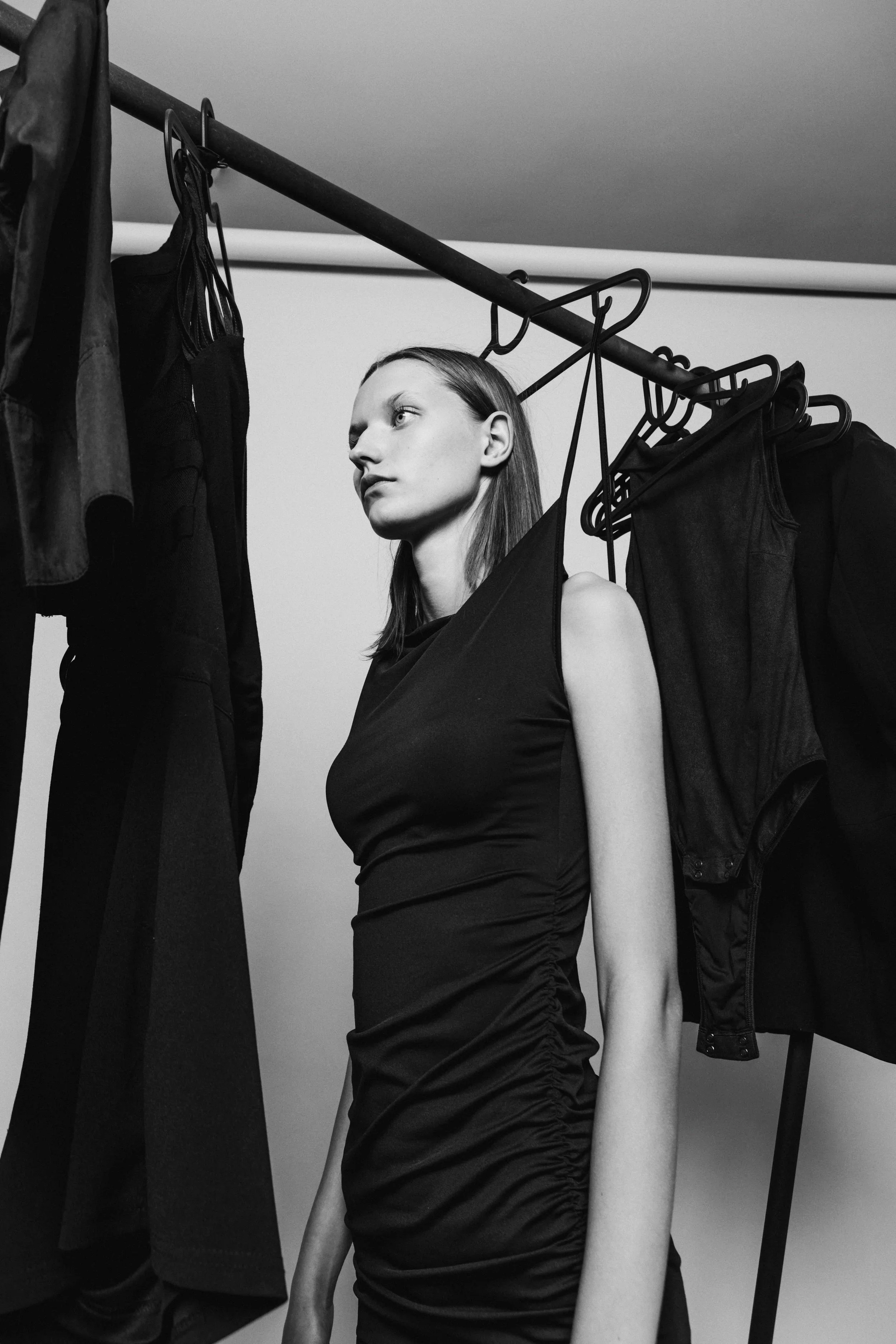 Black and white photo of a woman standing between hanging clothes, looking to the side with a serious expression.