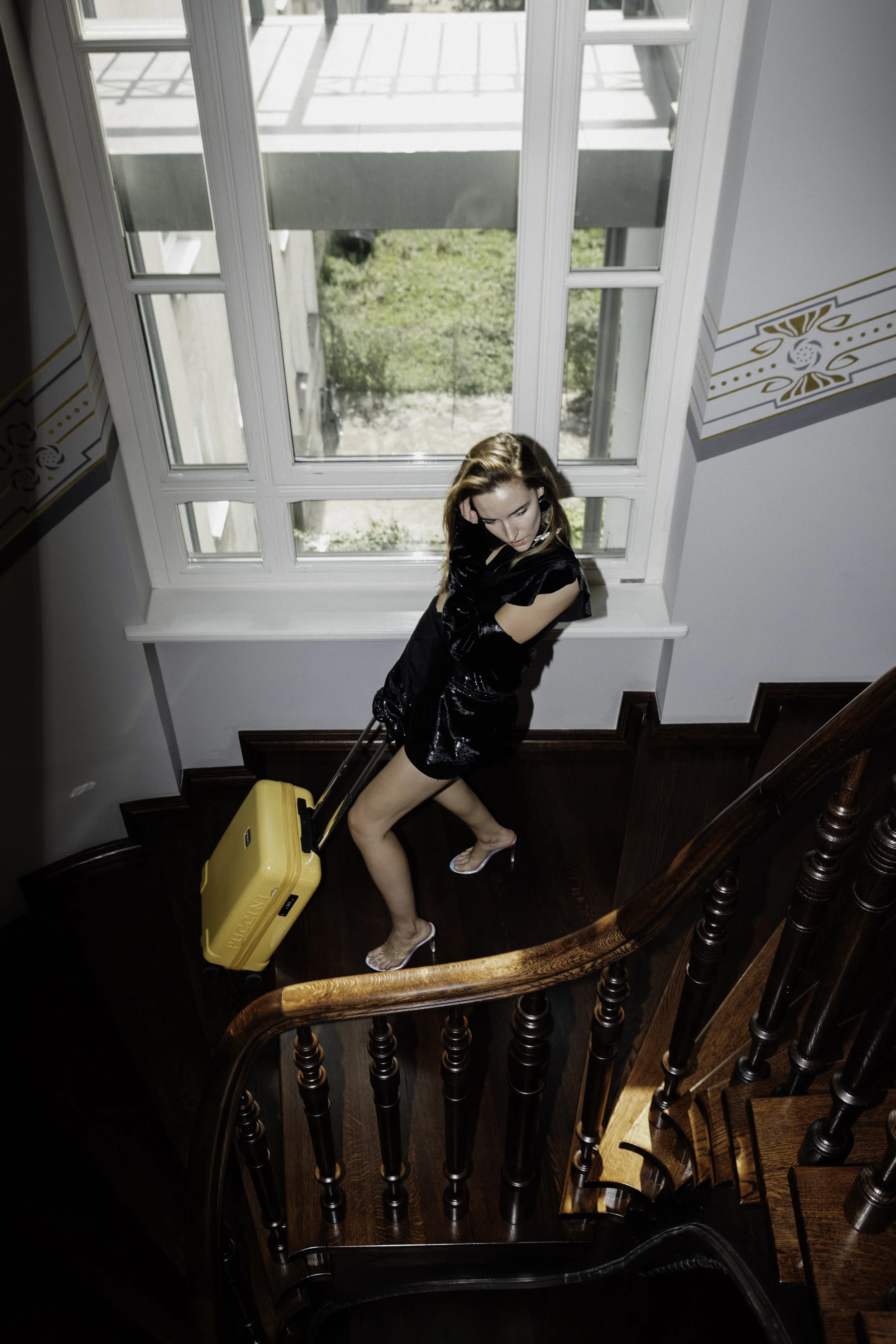 Woman in a black dress and heels walking up a wooden staircase with a yellow suitcase, near a large window showing a grassy outdoor area.