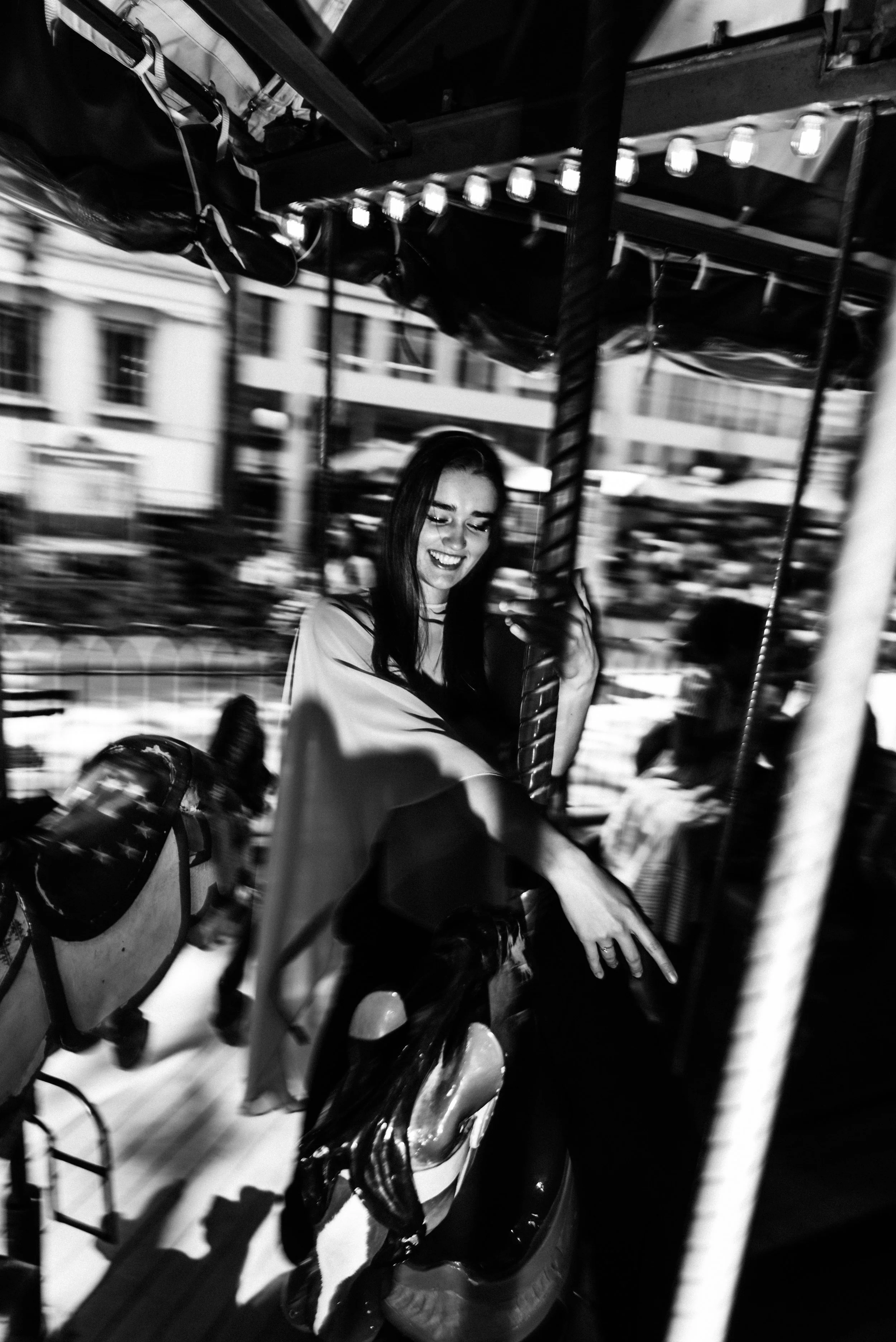 Black and white photo of a young woman laughing while riding a carousel horse outdoors.