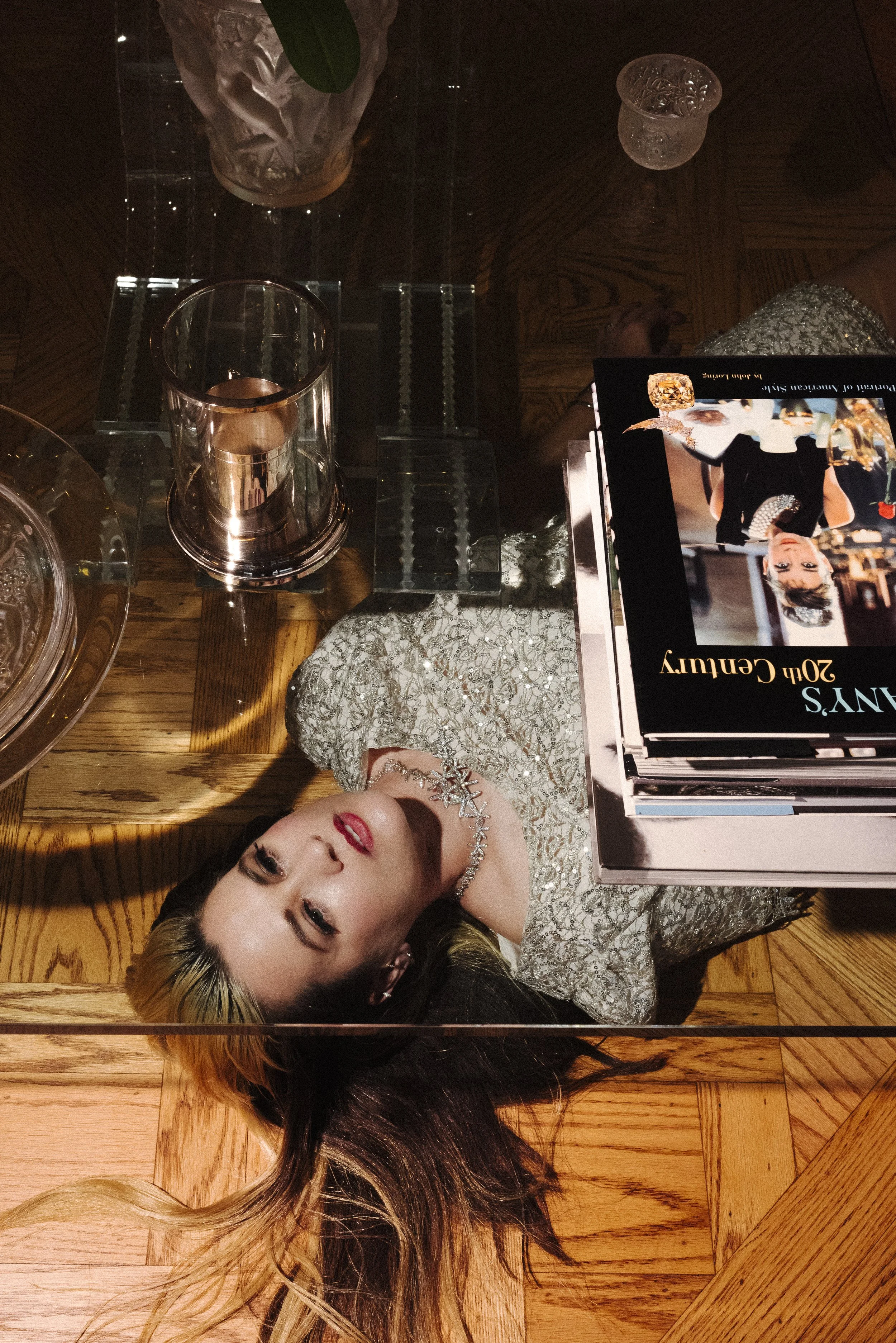 A woman lying on a wooden table, wearing a silver, beaded dress and a sparkling necklace, with hair spread out. There are glasses and books on the table, and the photo is taken from above, with a mirror reflection showing her face and upper body.