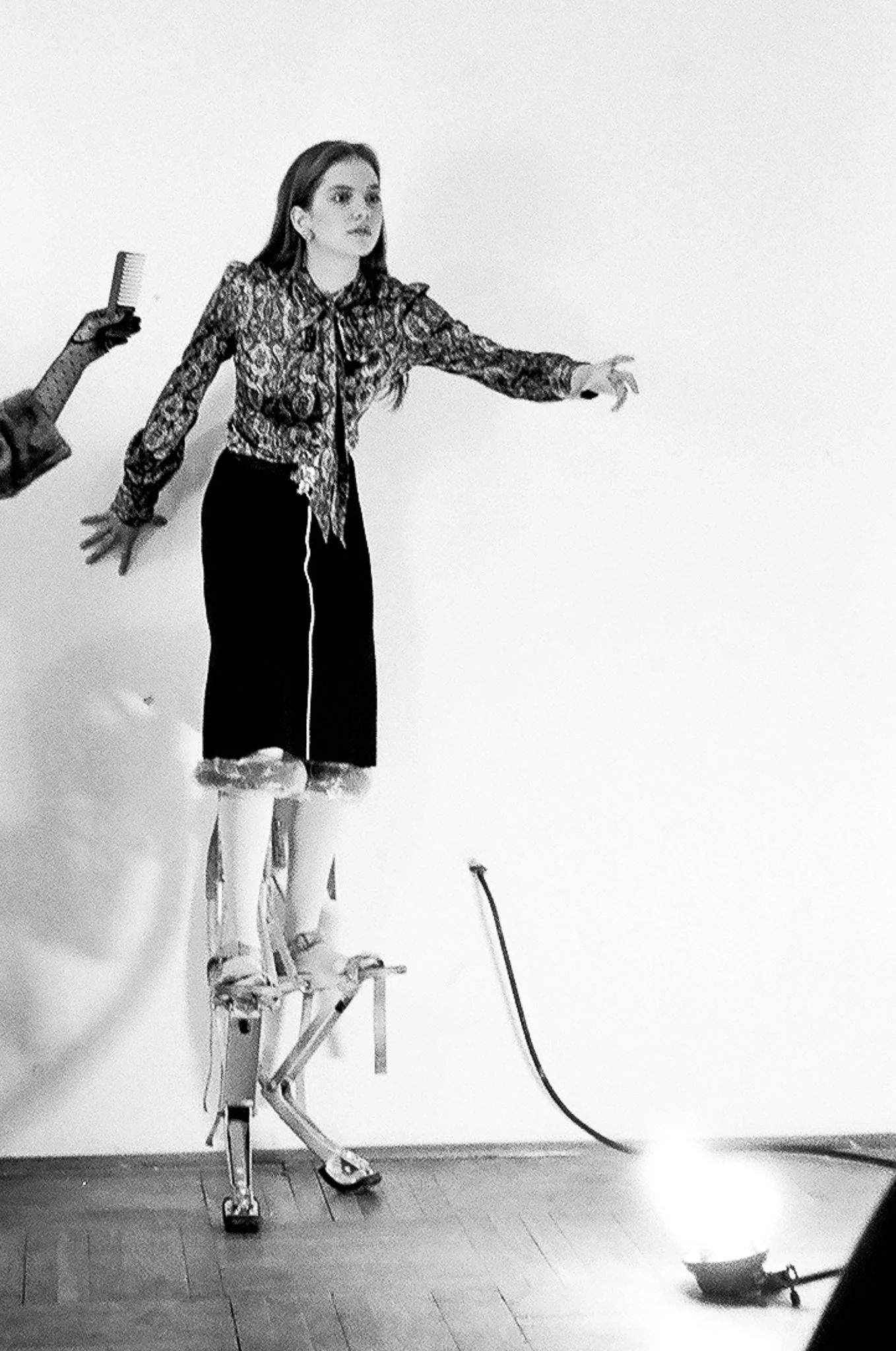 A woman is standing on a unicycle, reaching out with her right arm, in a room with a plain wall. There is a person partially visible holding a hairbrush behind her. A cord is hanging from her waist, and a small light or object is on the floor nearby.