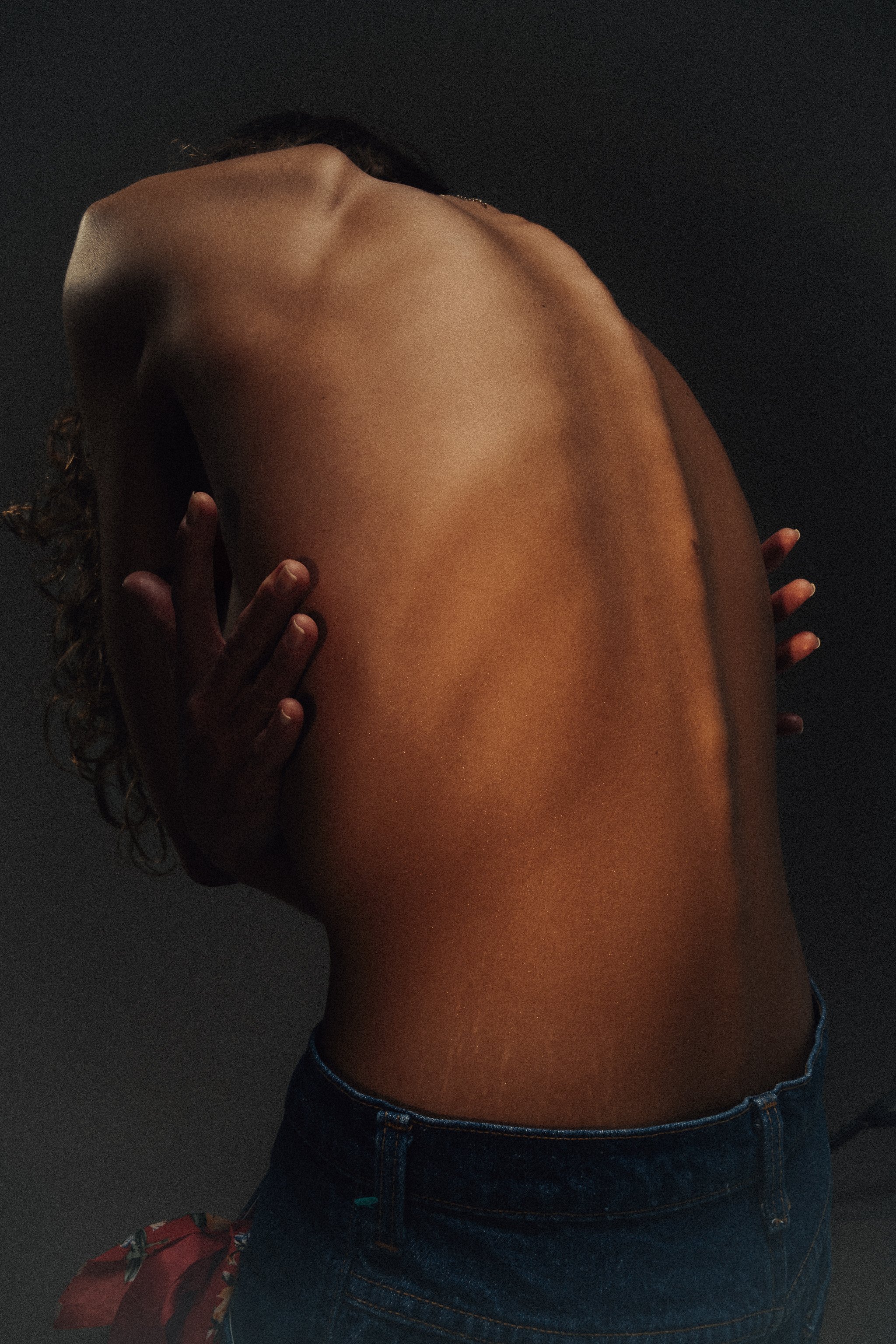A topless person with curly hair holding their arm across their chest, wearing jeans, against a dark background.