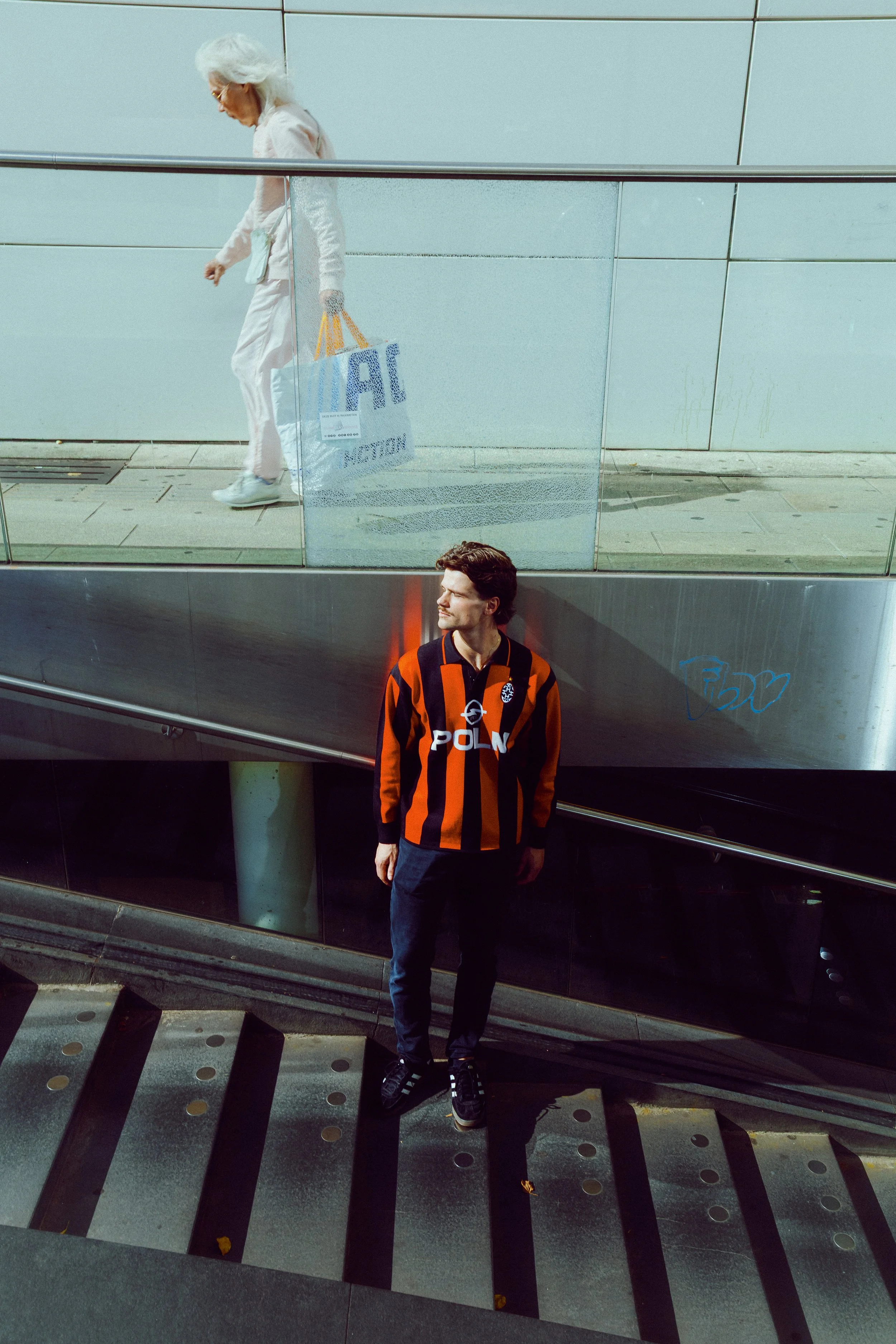 A young man with dark hair and light skin wearing an orange and black striped football jersey with 'POLAND' written across it, standing on a downward staircase at an airport. Above him, a reflection shows an elderly woman with white hair, dressed in 