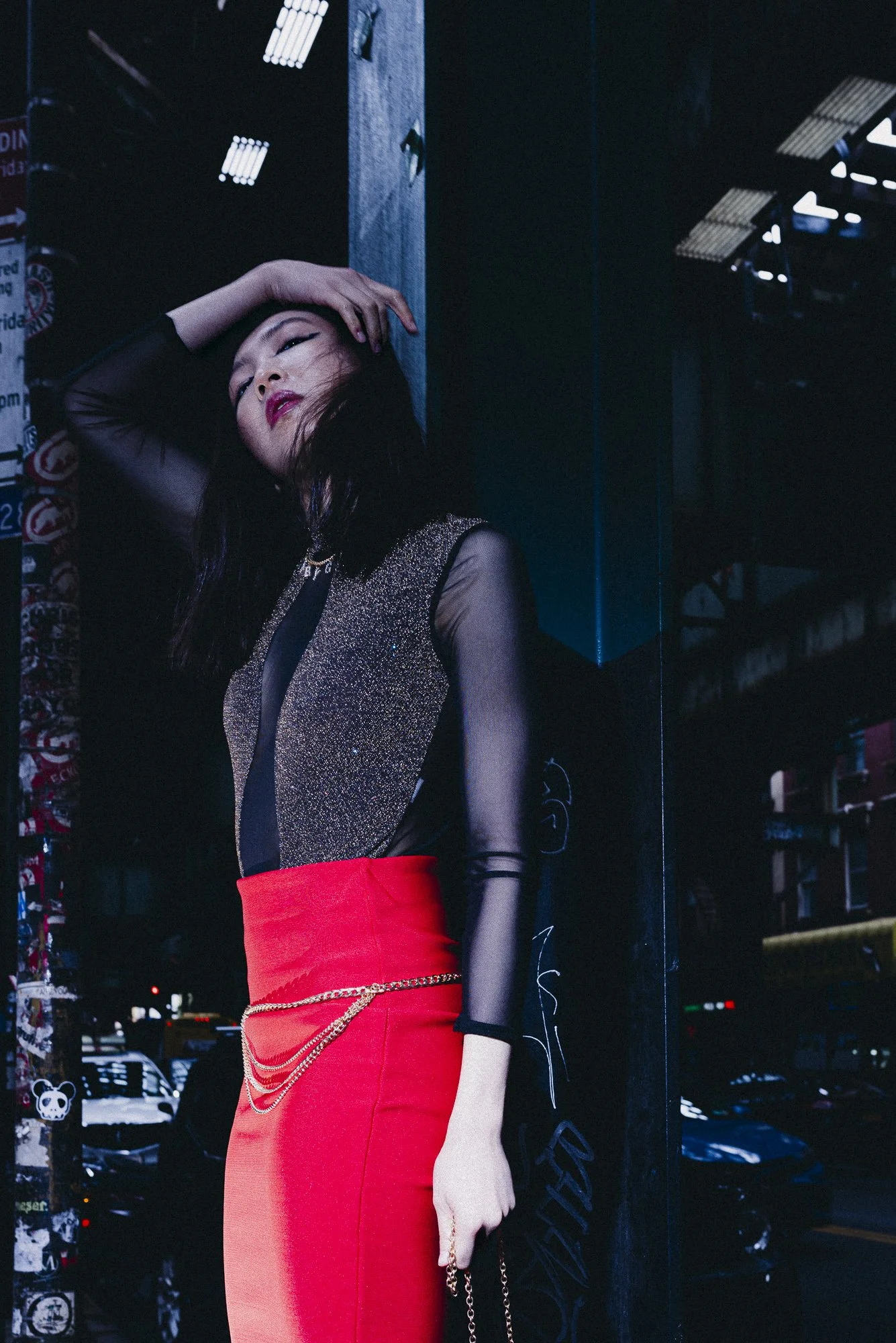 A woman with black hair and red lipstick leaning against a wall on a city street at night. She is wearing a black sheer top with gold accents, a red skirt with gold chains, and is posing with her head tilted back and one hand resting on her forehead.