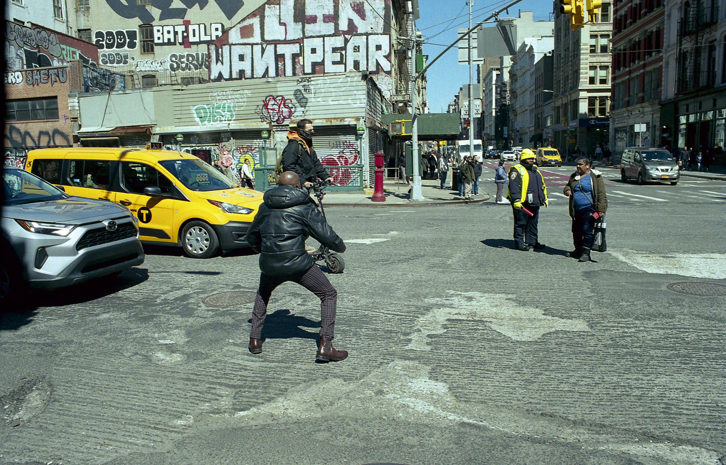 An urban street scene with a yellow taxi, a silver car, and several pedestrians crossing the intersection. The background features graffiti-covered buildings and storefronts.