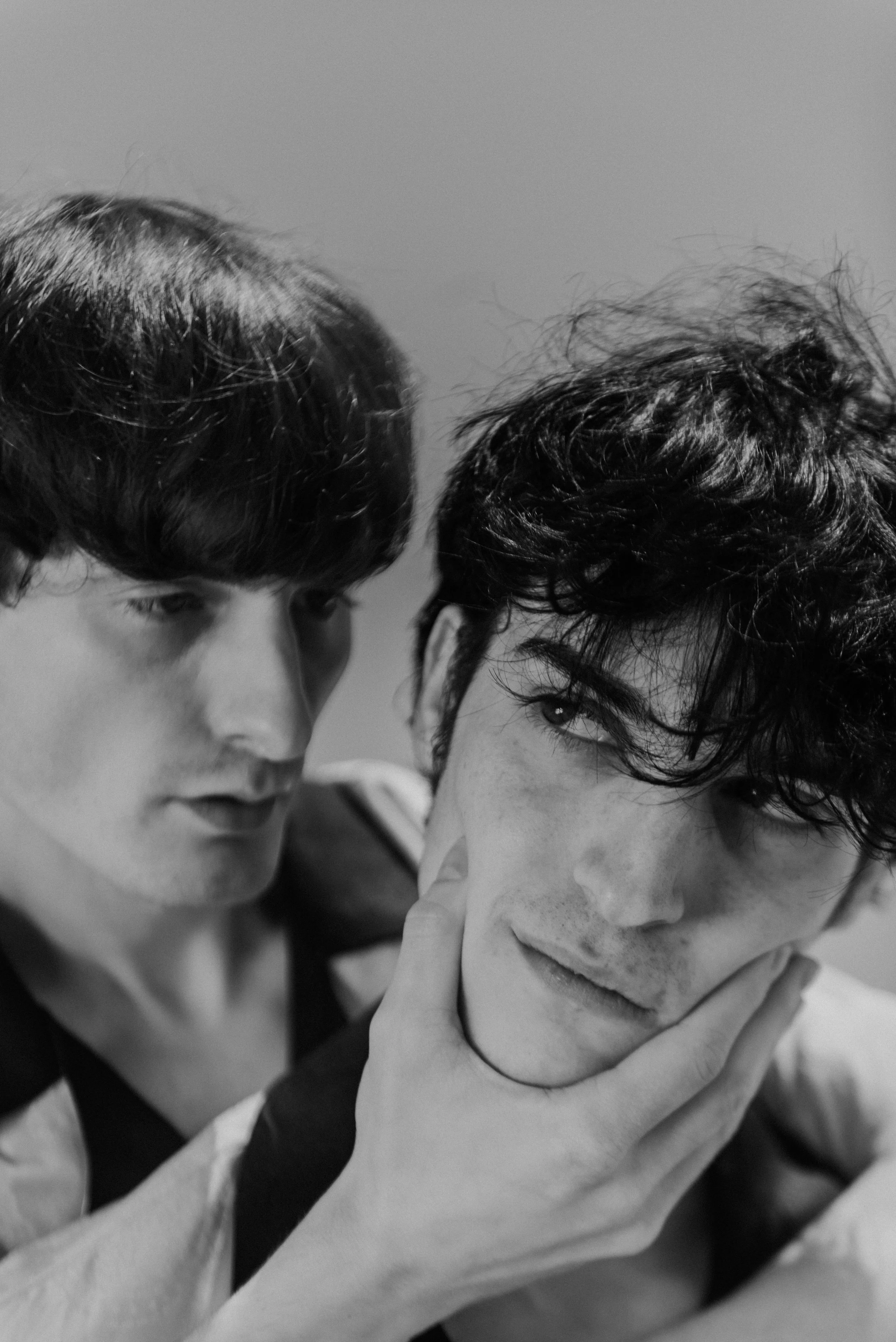 Two young men with dark curly hair, one gently holds the other's face, in a black and white close-up portrait.