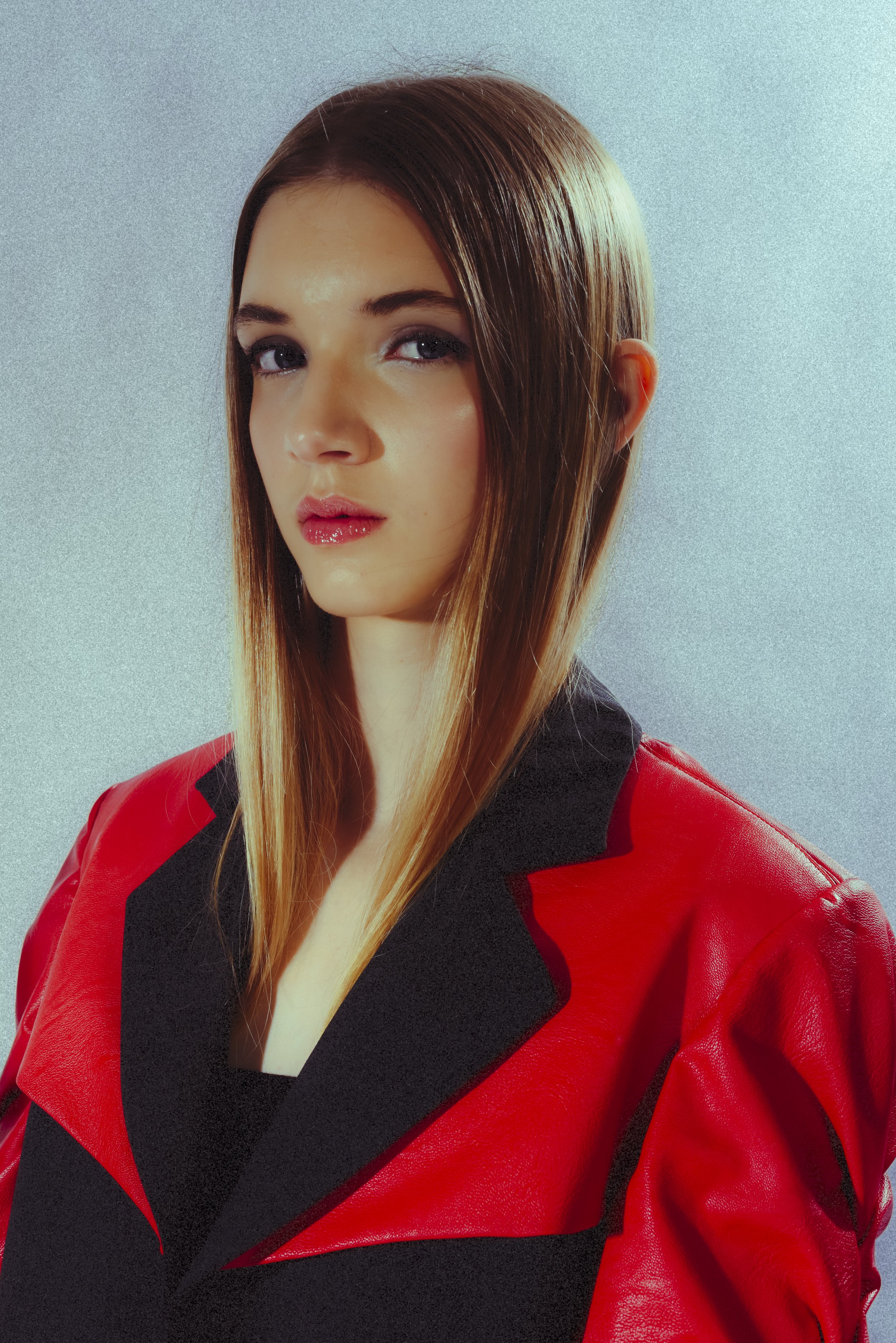 A woman with long straight brown hair wearing a red and black jacket looks at the camera against a light gray background.
