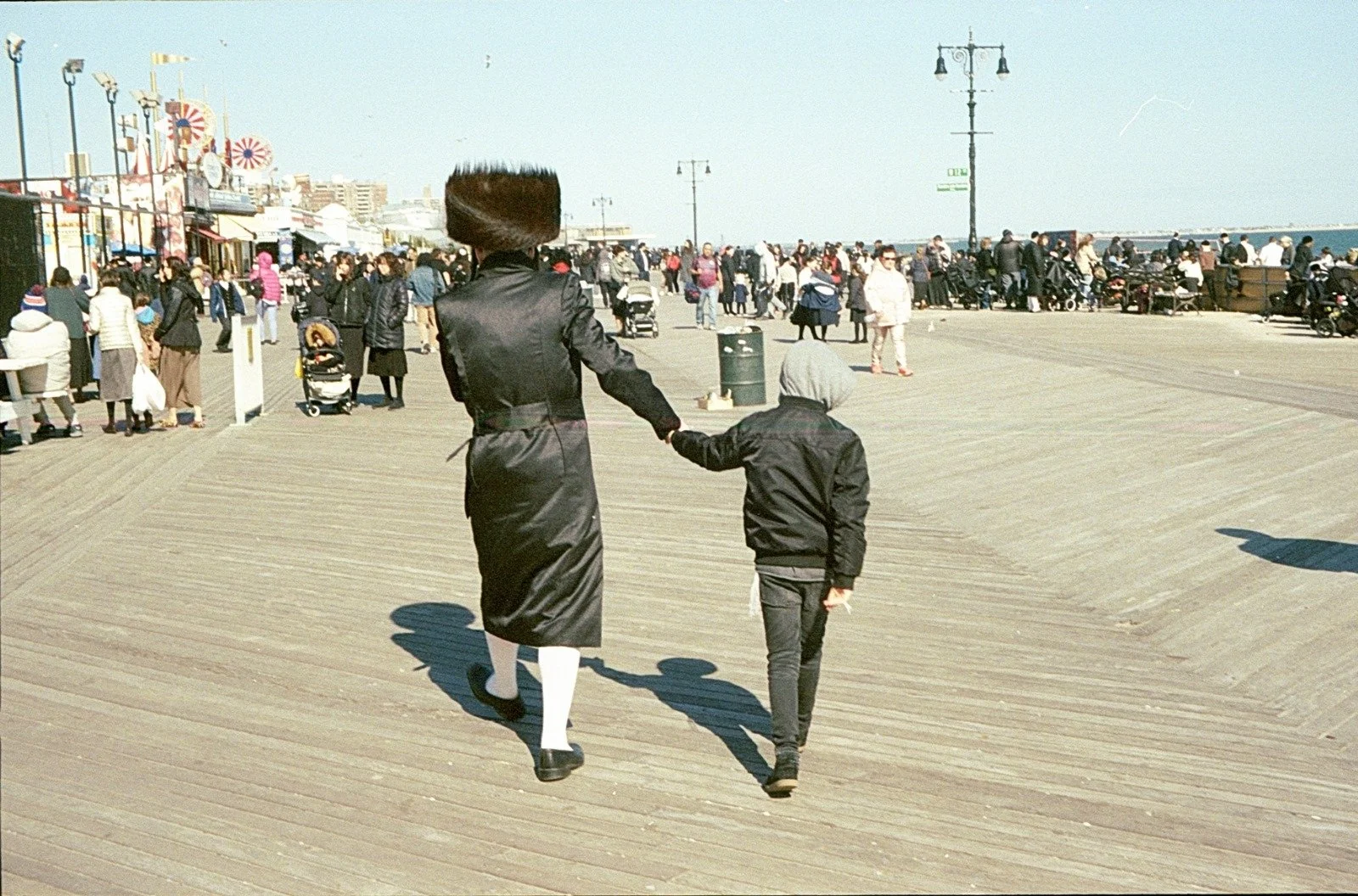 A woman and a child holding hands walk along a busy seaside boardwalk on a sunny day, with many people and amusement park rides visible in the background.