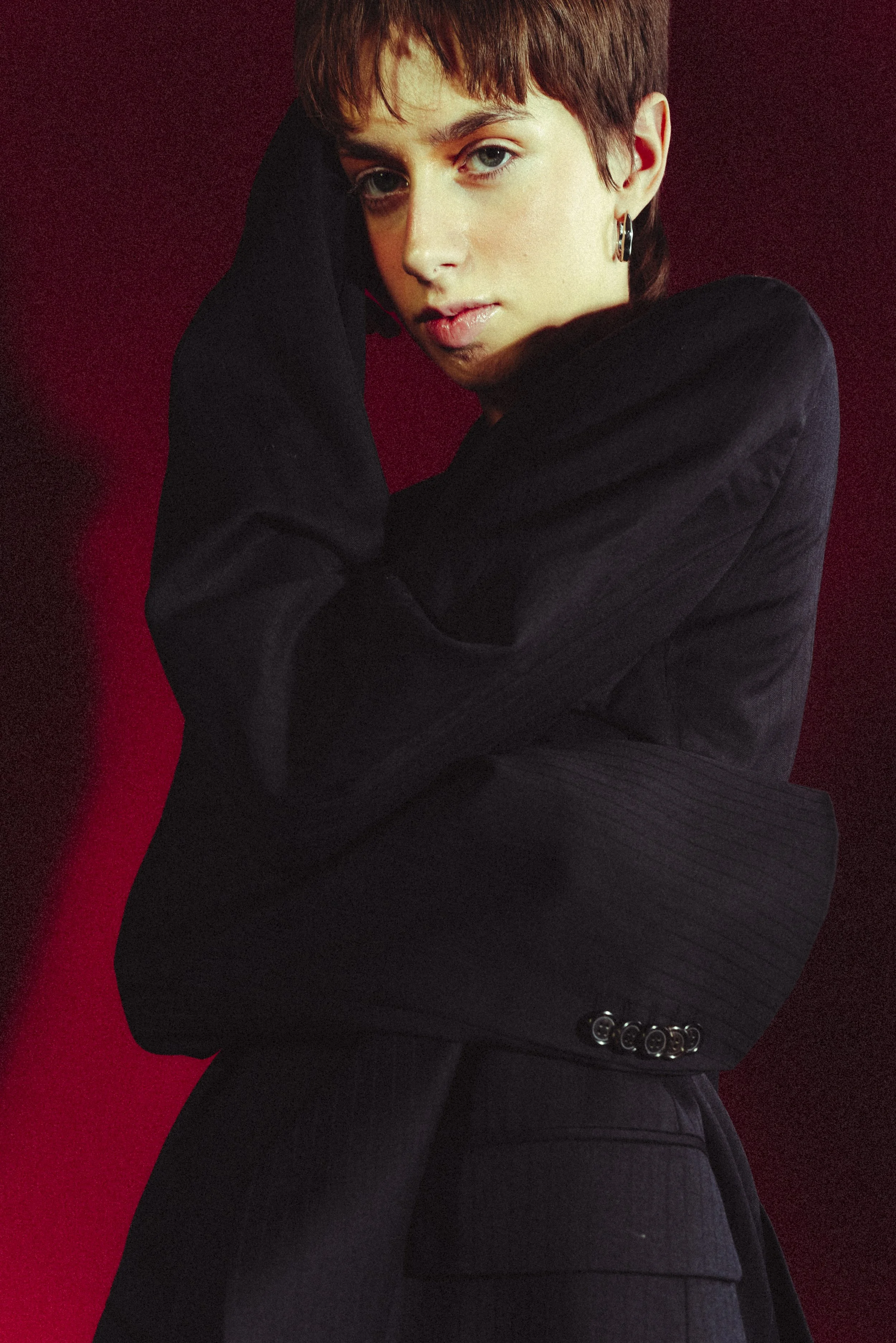 Young woman with short brown hair, wearing black clothing, posing against a red background.