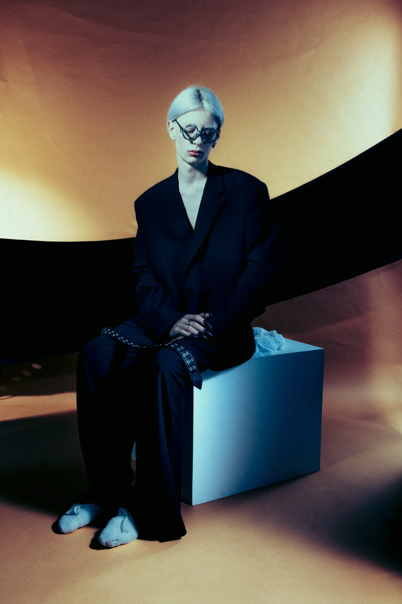 Person with platinum blonde hair and glasses, dressed in black, sitting on a white block against an abstract, colorful background.