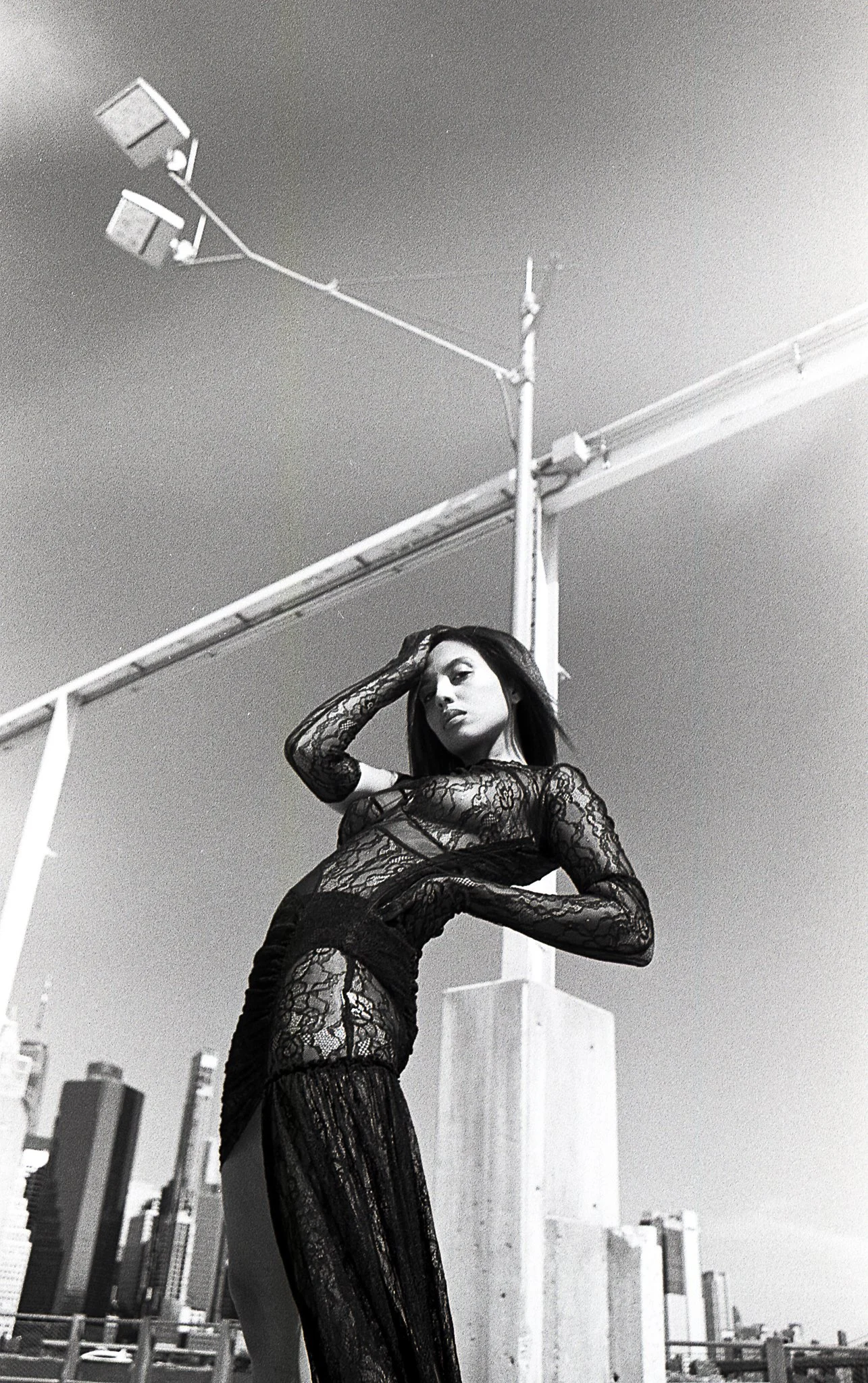 Black and white photo of a woman in a sheer, lace dress standing on a rooftop with city buildings in the background and a streetlight hanging from above.