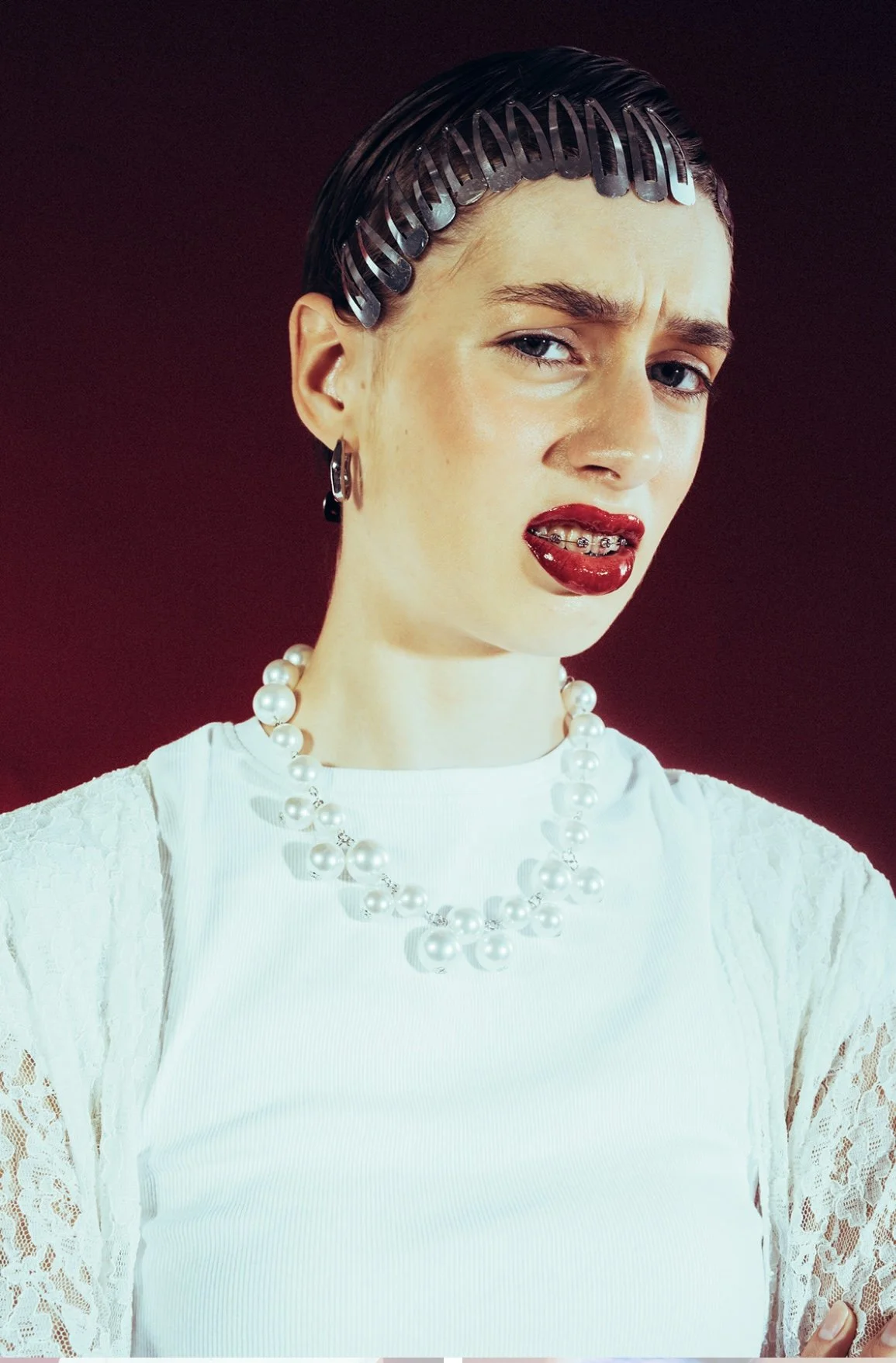 A young woman with light skin, dark hair held back with plastic hair clips, wearing a white lace top, pearl necklace, and earrings, making a confused or skeptical facial expression with her tongue slightly out.