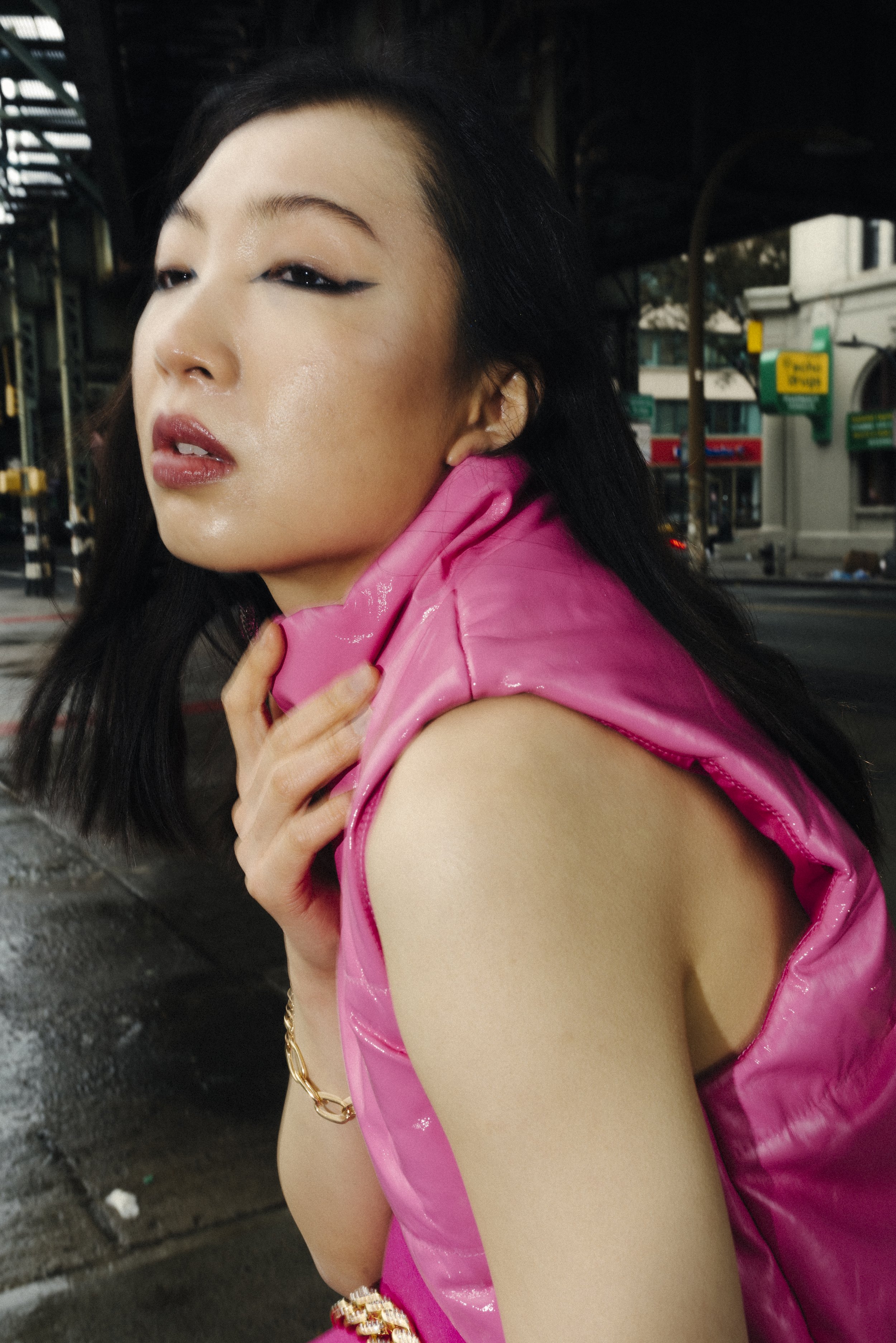 A woman with dark hair and makeup, wearing a pink shiny sleeveless jacket, is holding her shoulder in an urban street setting with store signs visible in the background.