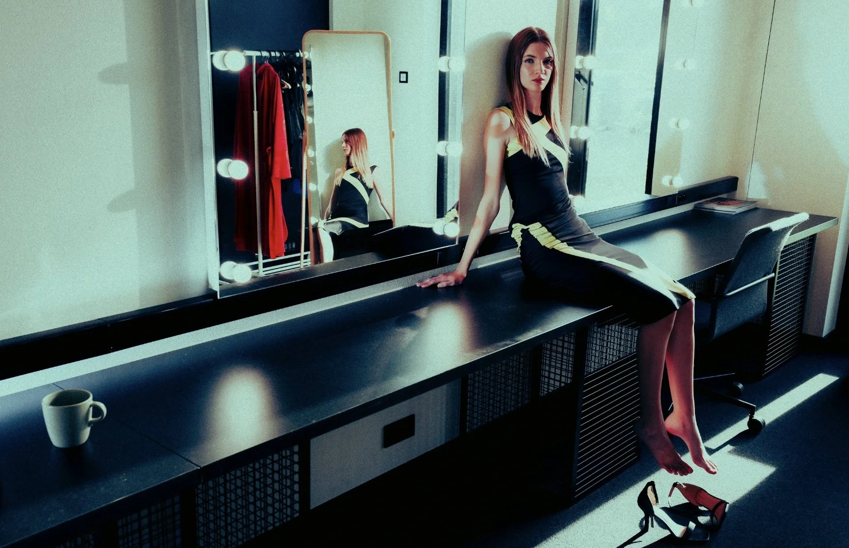 A woman with long red hair sitting on a black dressing table in front of a mirror with bright lights. The mirror reflects her and some red and black clothing. A cup is on the table, and red and black shoes are on the floor. She is wearing a black and