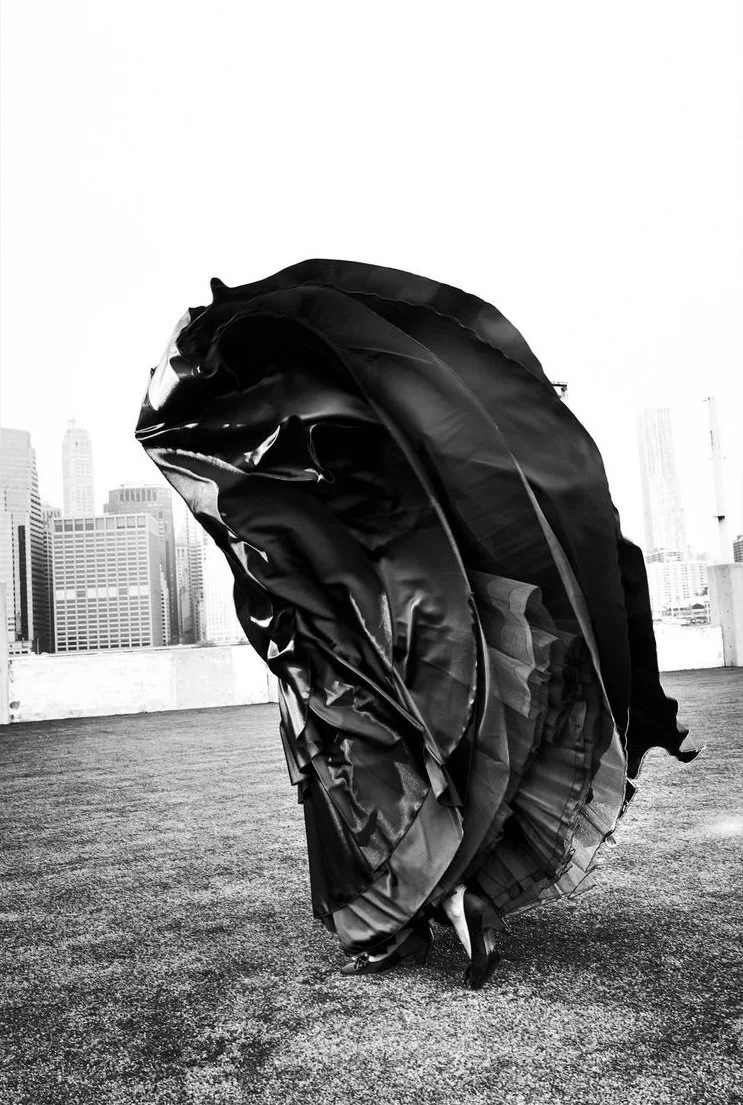 Person dressed in a black gown with large, flowing fabric, standing on a rooftop with a city skyline in the background.