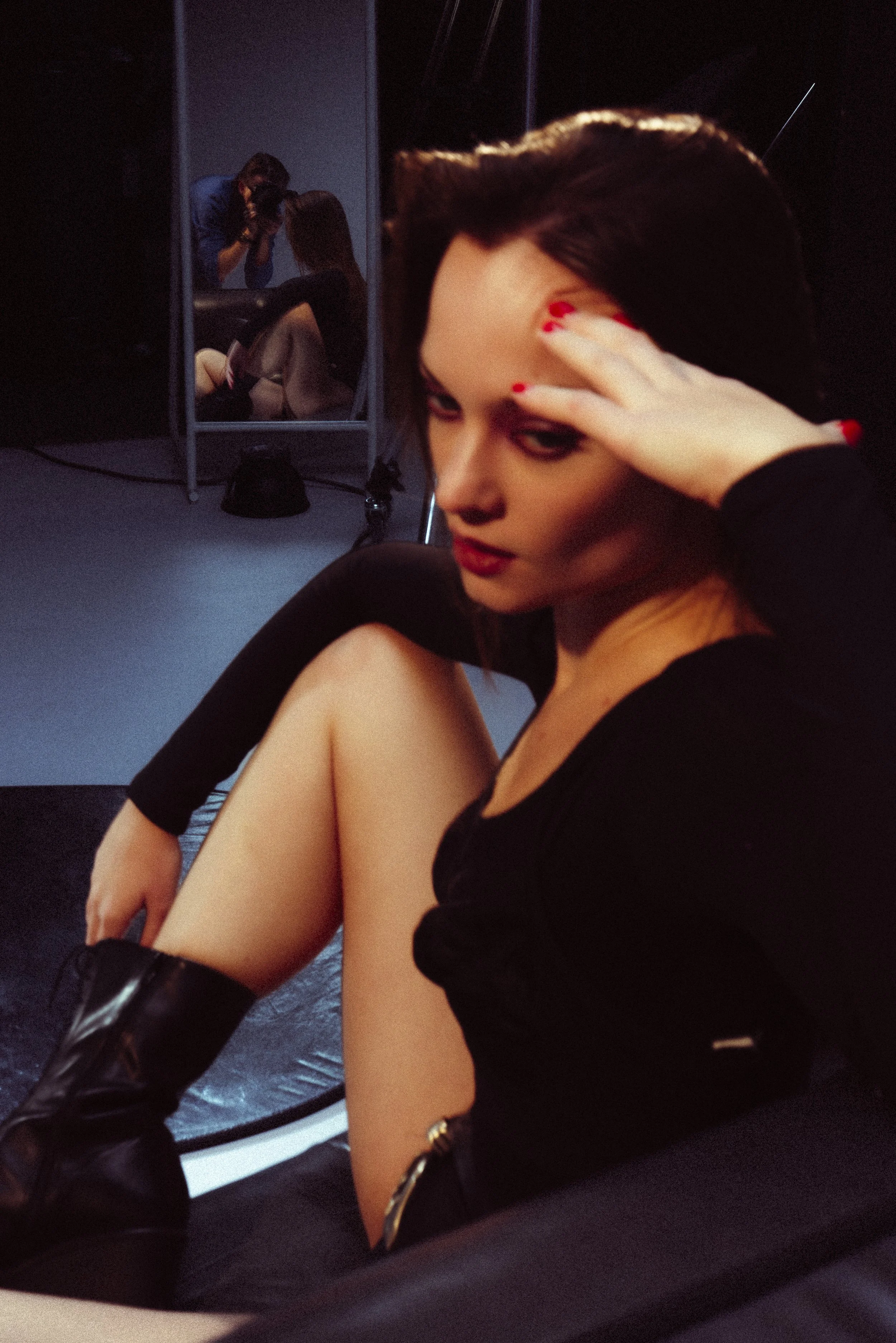 A young woman with red nail polish sitting on a dark floor, wearing a black outfit and black boots. She has dark hair and is looking at the camera with her hand on her forehead. A mirror behind her reflects her and a photographer taking her picture i