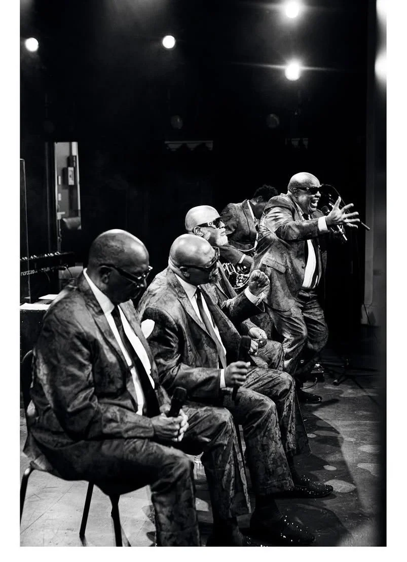Five men dressed in suits and sunglasses sitting and standing on stage during a performance or event. One man standing, singing or speaking passionately with a microphone in hand. The scene is in black and white.