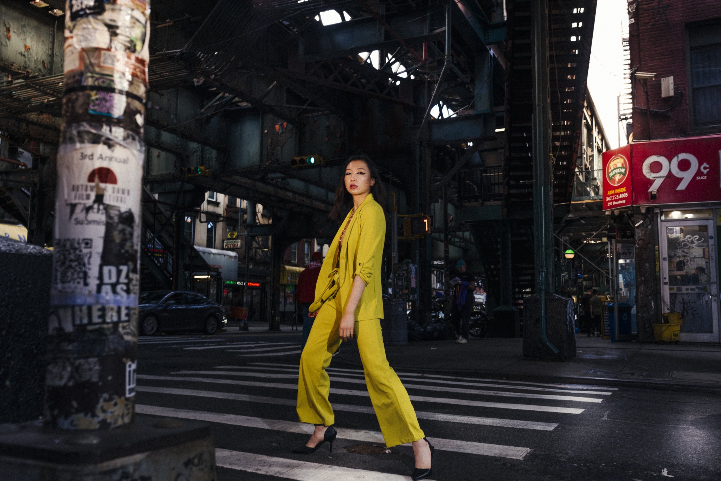 A woman in a bright yellow suit and black high heels crossing a city street at night under an elevated train track, with cars and pedestrians in the background.