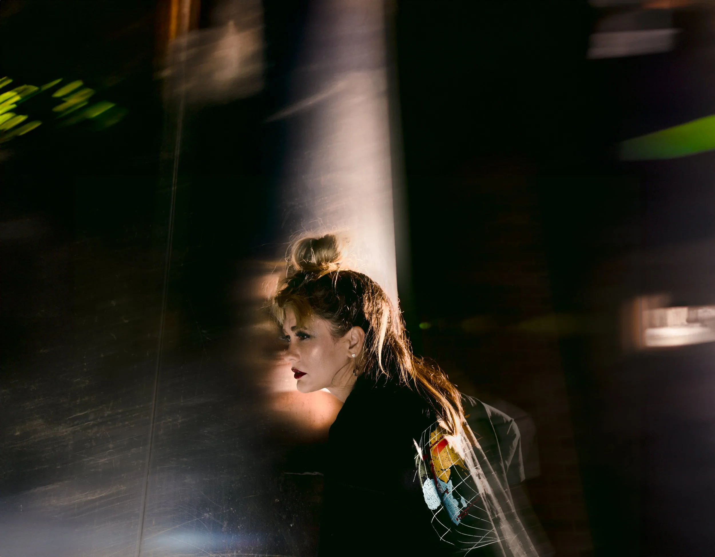A young woman with long hair tied in a bun leaning against a reflective metallic wall, illuminated by warm light, with a side profile and serious expression, wearing a jacket with a colorful pattern on the sleeve.