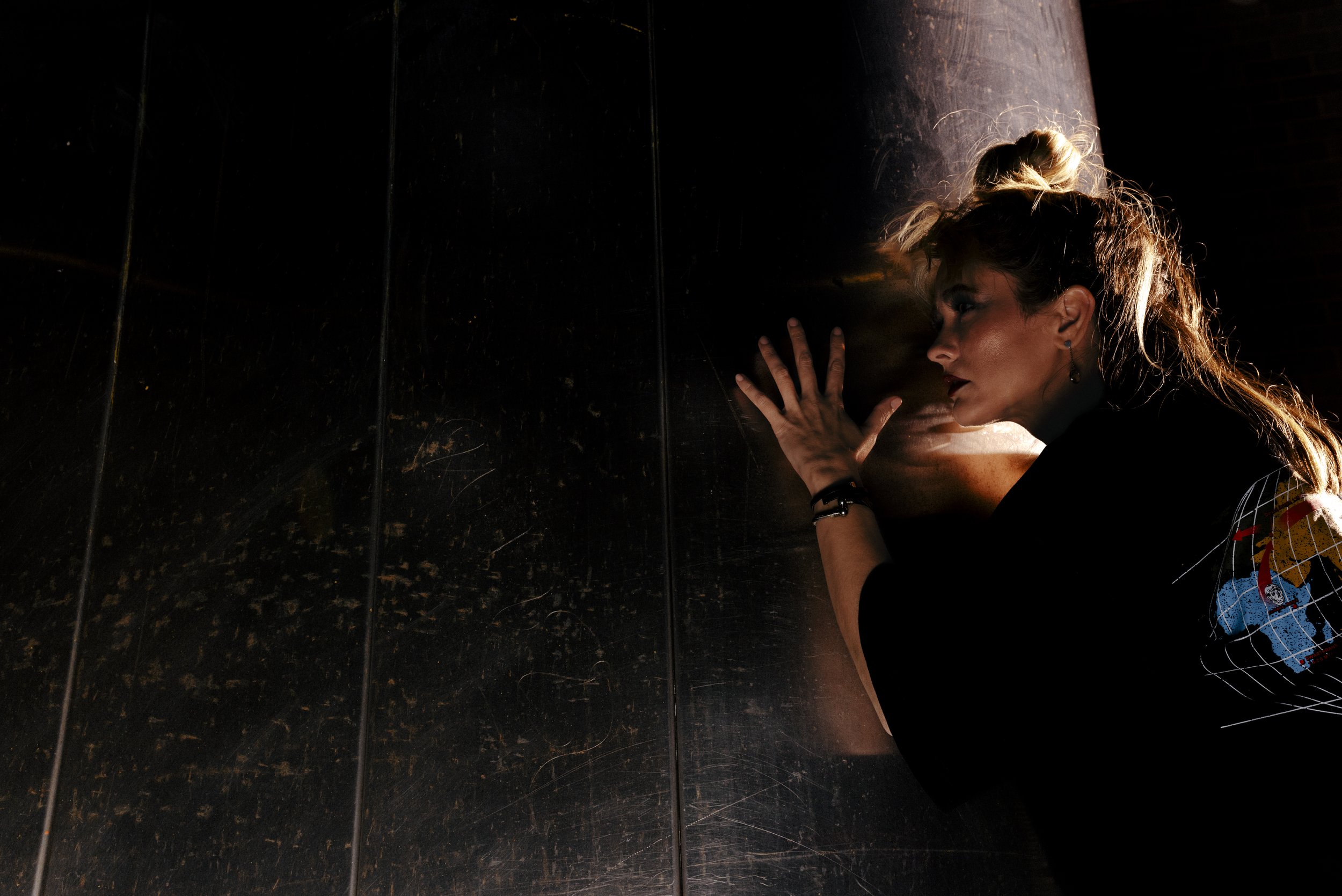 A woman with blonde hair in a ponytail, wearing dark clothing and earrings, leaning her head and hand against a dark mirrored wall, with her eyes closed and lips slightly parted.