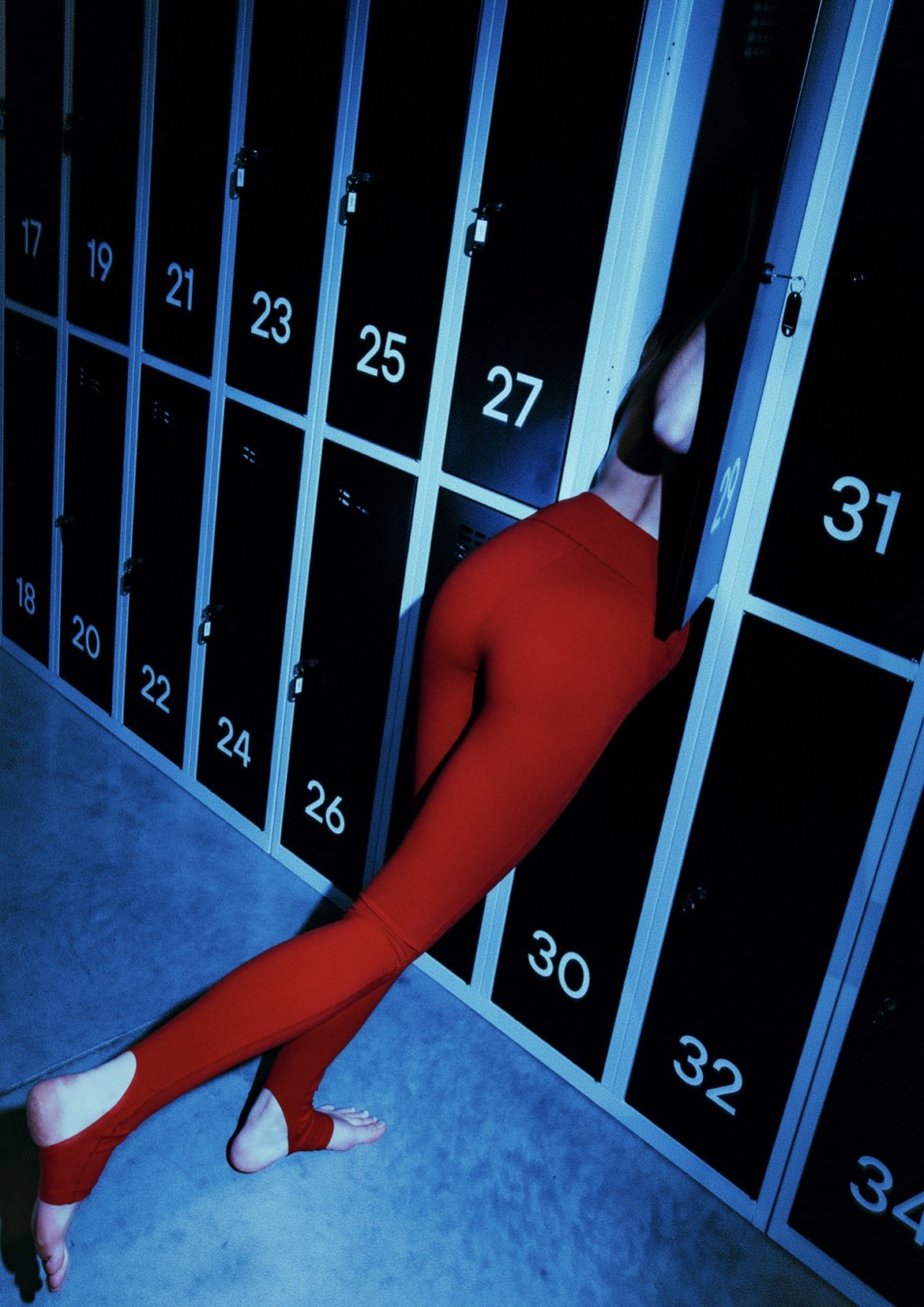 A person in red leggings is leaning into a locker, with only part of their body visible, in a locker room with numbered lockers.