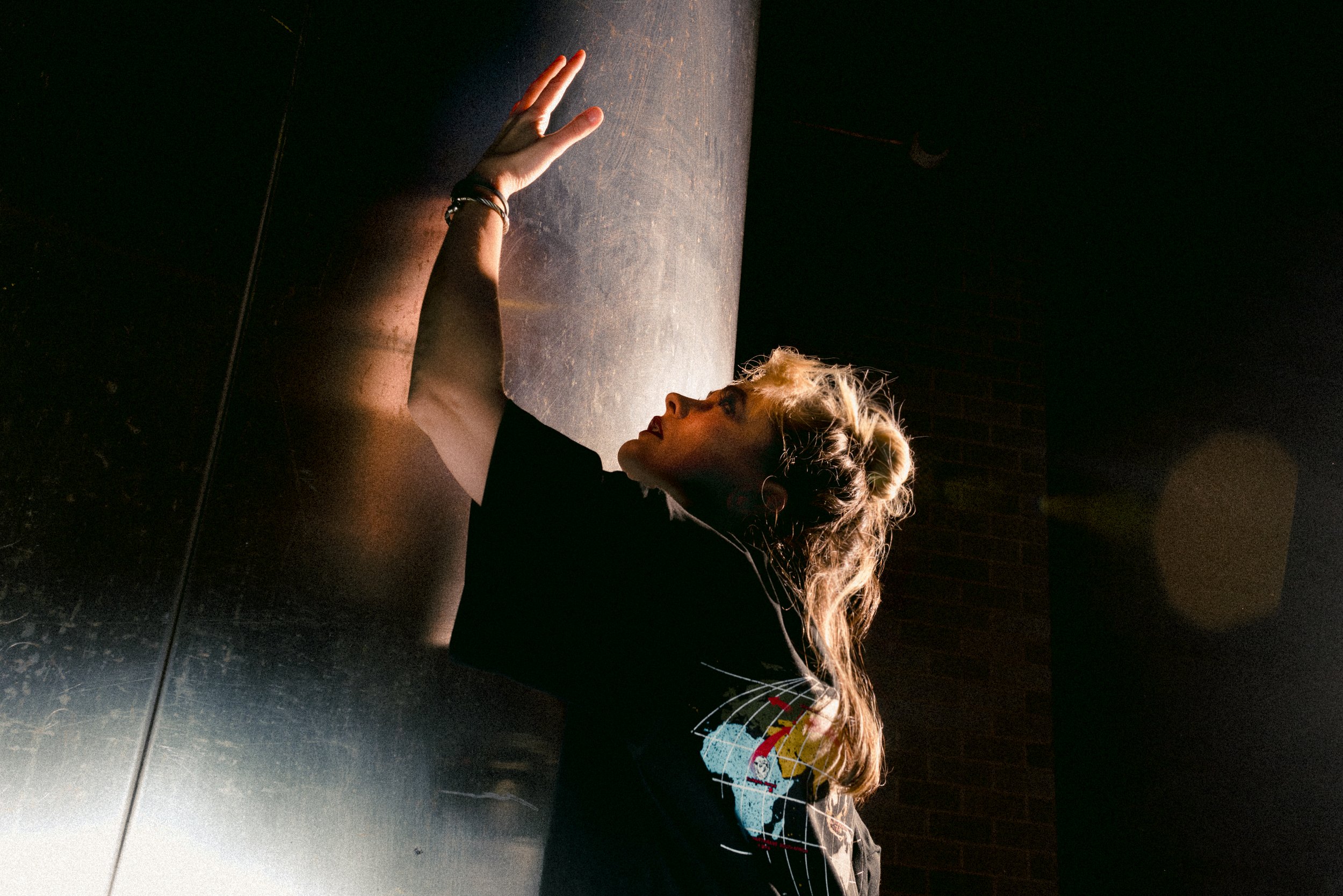A woman with blonde hair tied in a bun, wearing a black shirt with colorful graphic, reaches up with her left hand, touching a vertical surface in a dark room with light illuminating her face and arm.