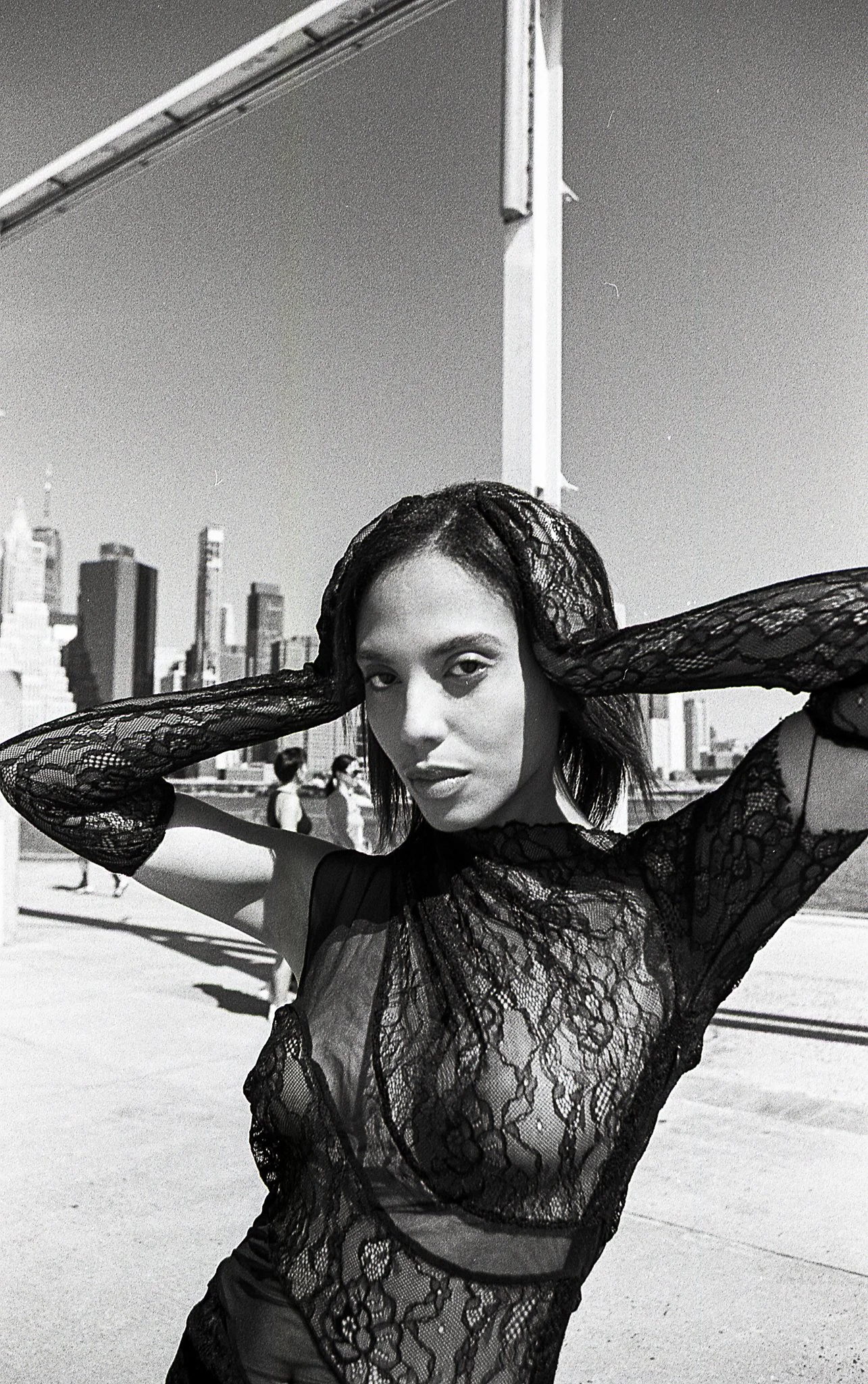 A woman with short hair and dark lace top posing outdoors in front of a city skyline, with her hands behind her head.