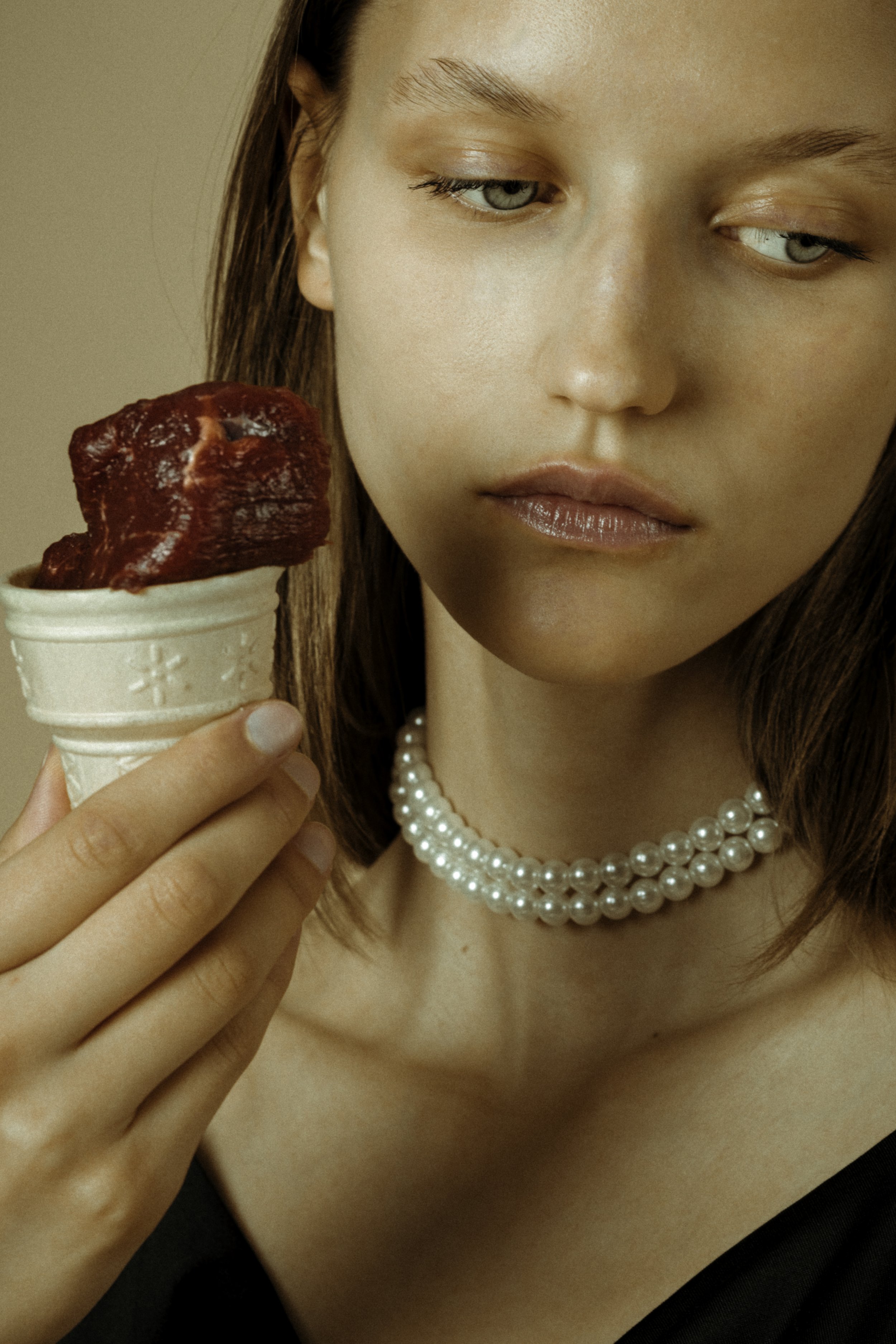 A woman with brown hair and pearl necklace holding an ice cream cone with red sorbet, looking at it with a neutral expression.