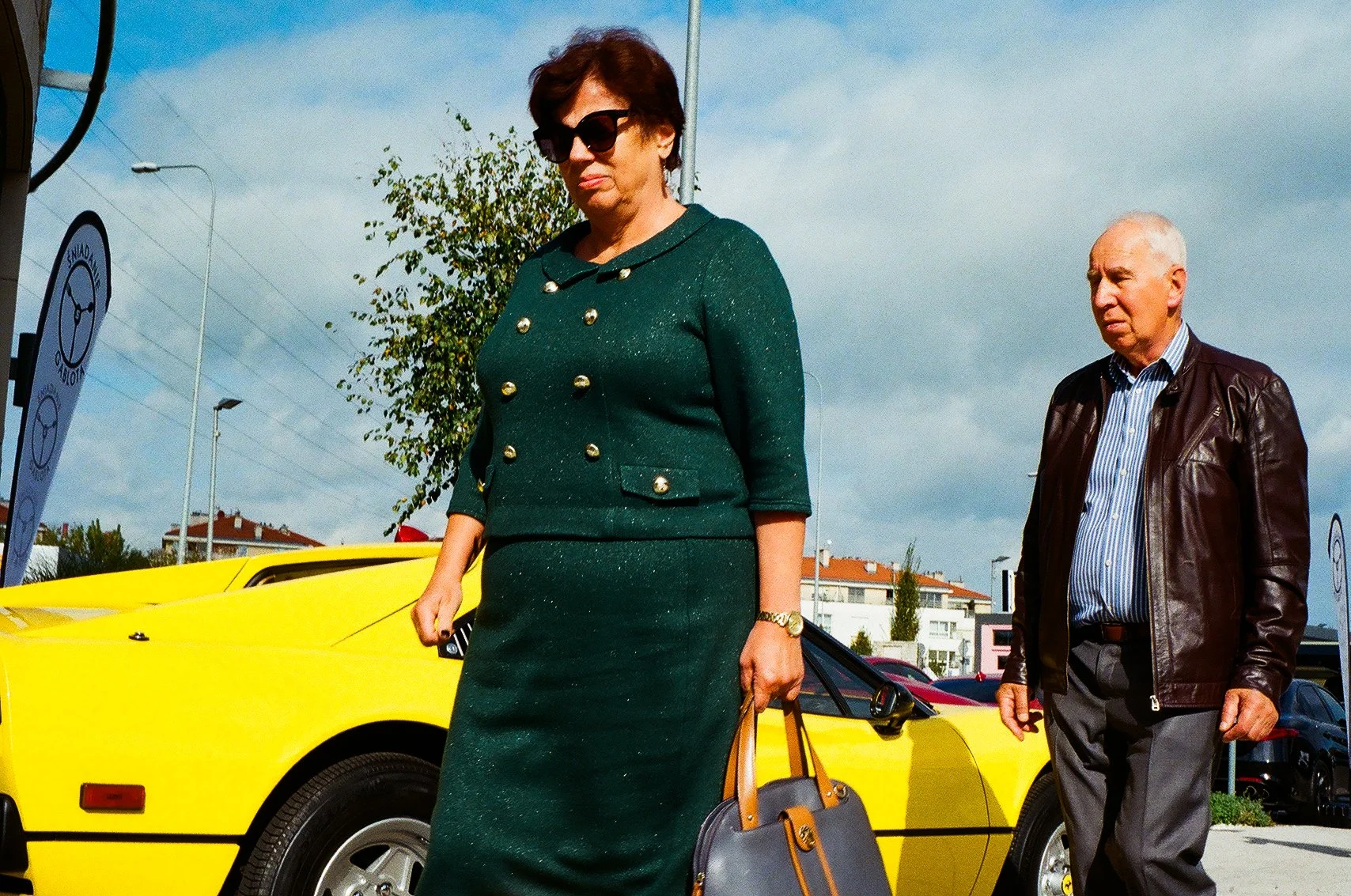 A woman wearing sunglasses and a green dress with gold buttons walks past a yellow sports car. An older man in a leather jacket and striped shirt walks nearby in the parking lot under a cloudy sky.