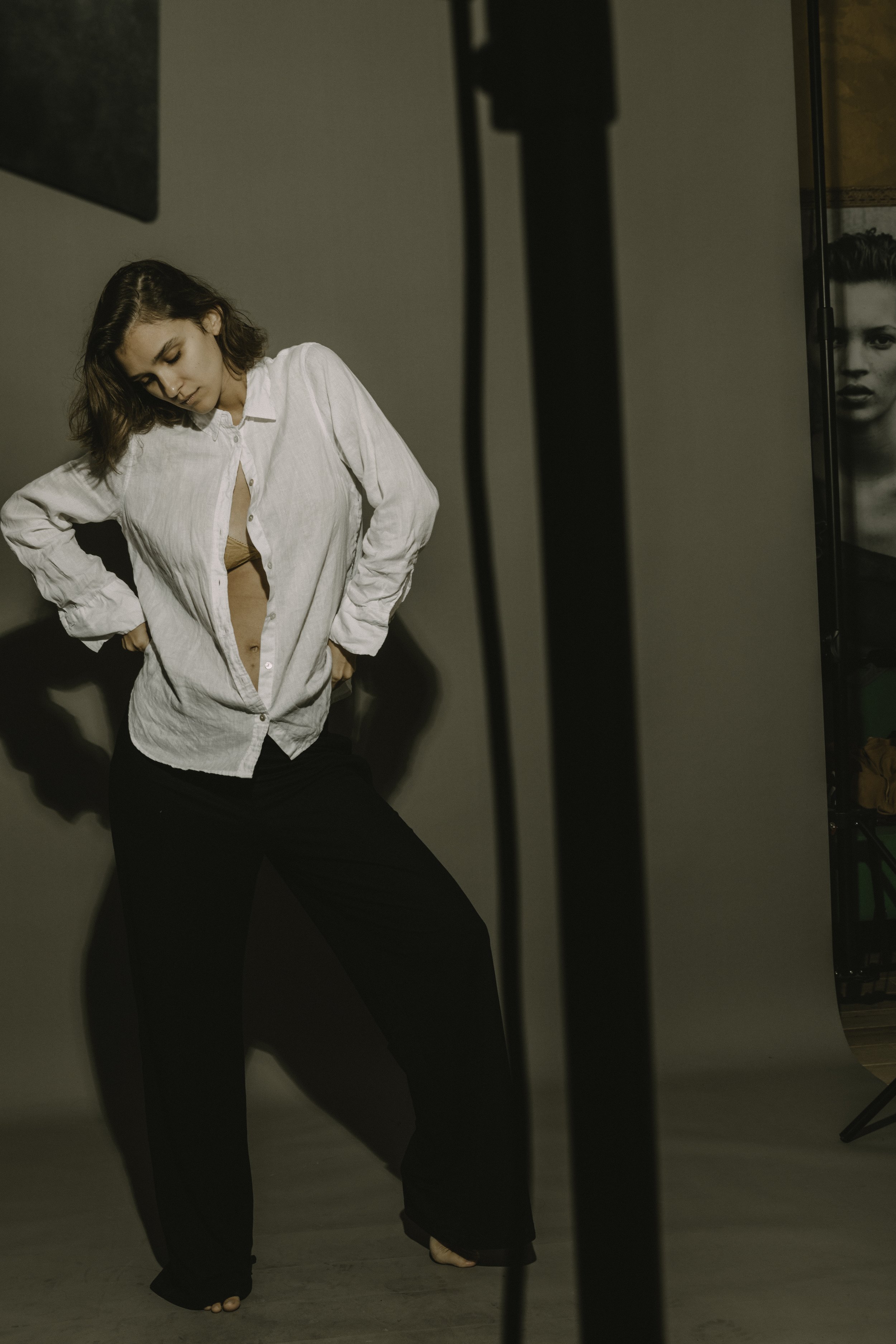 A woman with short brown hair wearing a white shirt and black pants standing barefoot on a brown floor, with her head tilted down and her hands on her hips, in a photography studio setting.