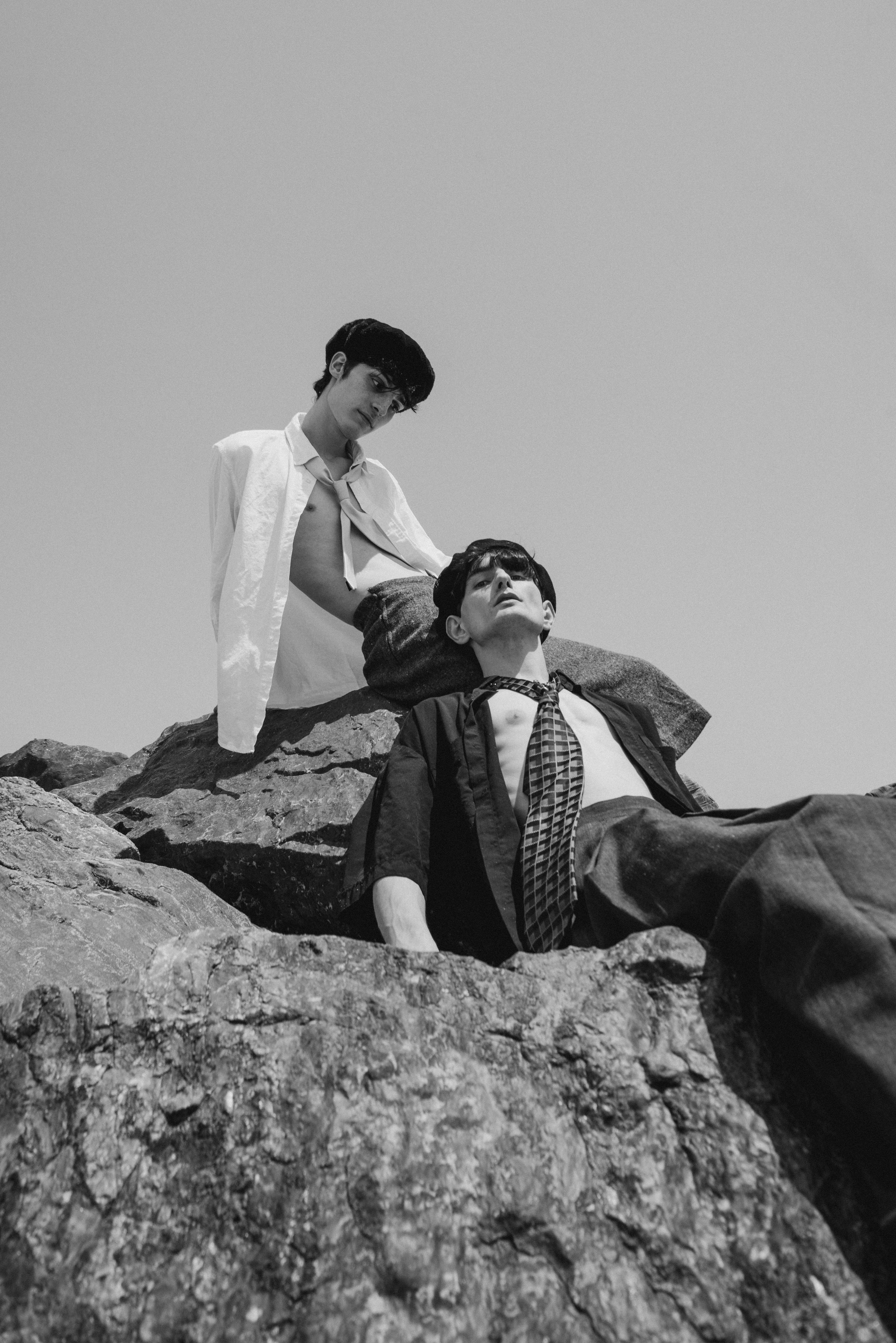 Two young men with dark hair and slim builds, one sitting and the other reclining on rocks, in black and white, wearing casual and formal clothing respectively, under a clear sky.