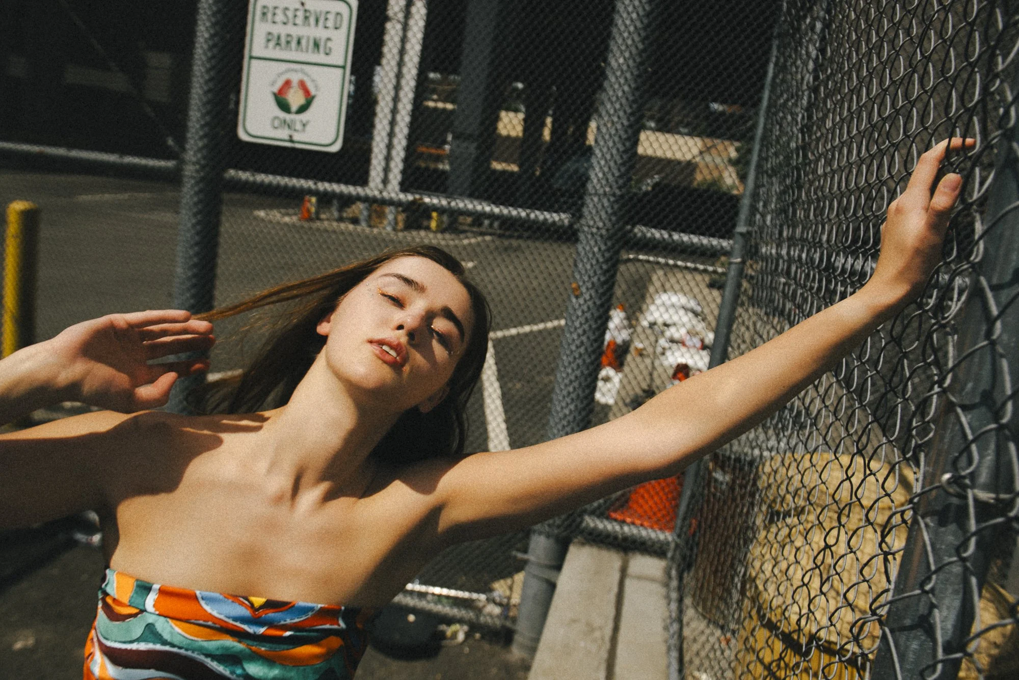A woman wearing a colorful strapless dress leaning against a chain-link fence in an outdoor parking lot.
