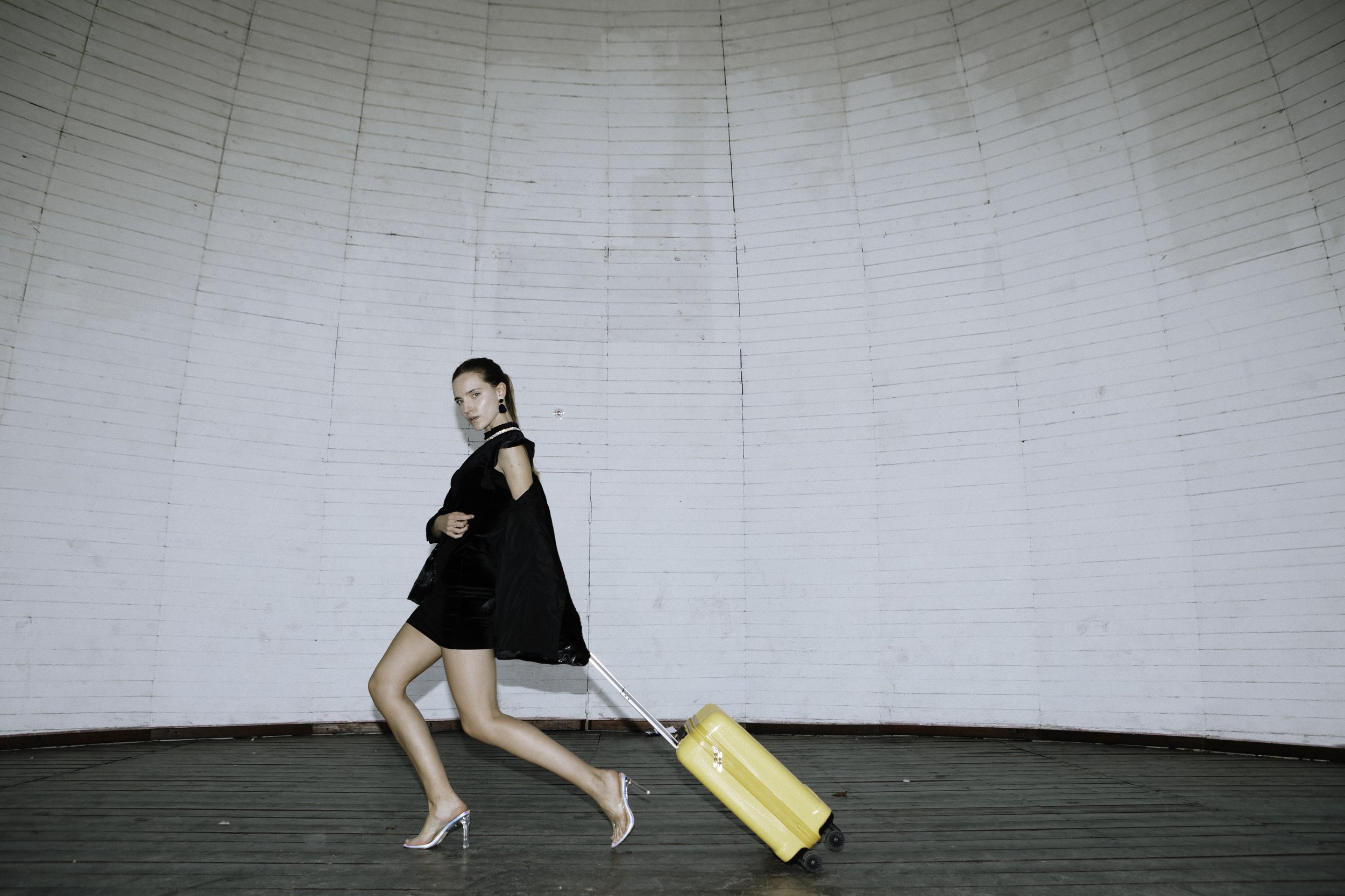 A woman in high heels and a black outfit, standing on a wooden platform, pulling a yellow rolling suitcase that is leaning back on its wheels. She stands in front of a large, gray, semi-circular wall.