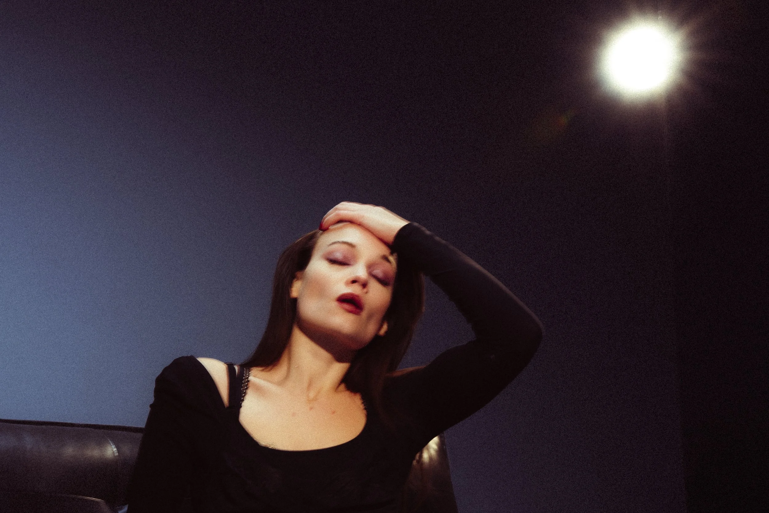 A woman with dark hair and makeup posing with her hand on her head, sitting against a dark blue wall with a bright light in the upper right corner.