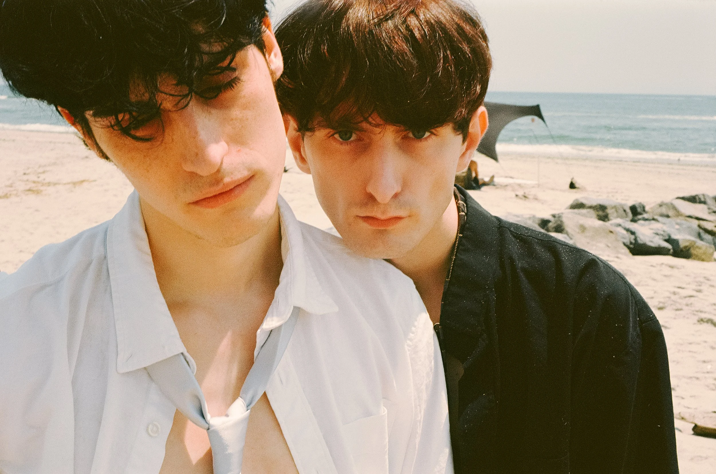 Two young men standing close together on a beach, looking into the camera, with rocks, sand, and the ocean in the background.