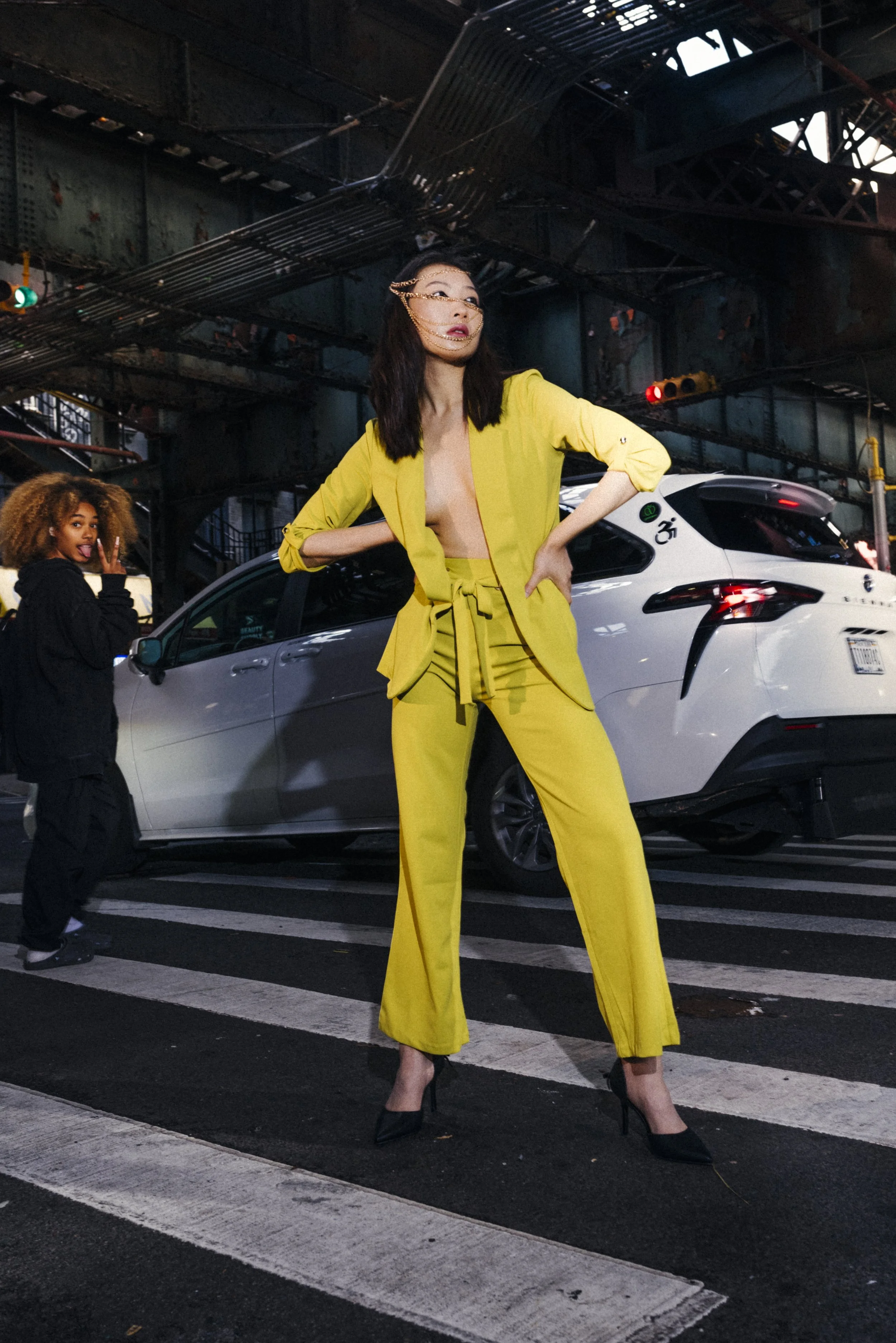 A woman in a yellow pantsuit and high heels stands confidently in the middle of a city street at night, with a silver car behind her and another woman in black to the side, under a bridge or elevated structure.