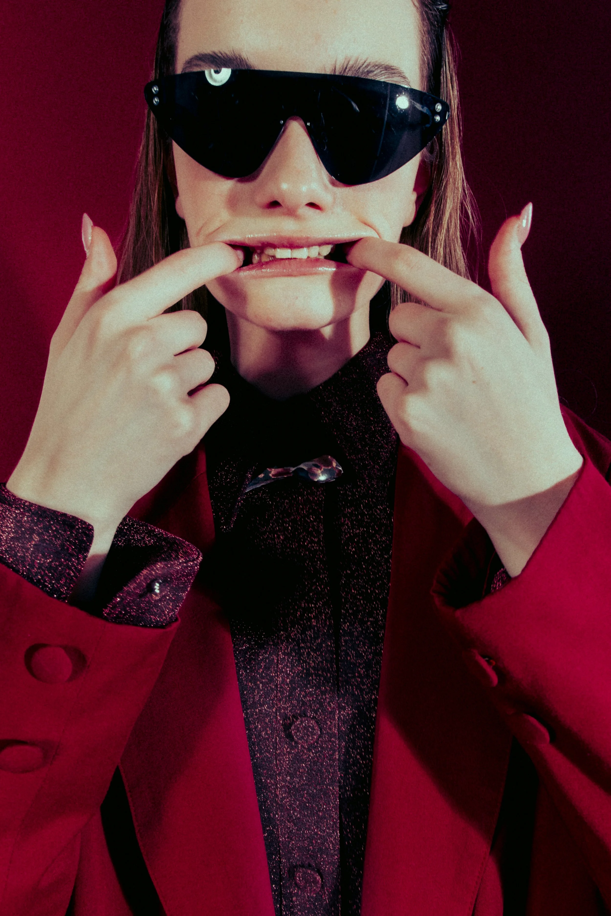 A woman wearing black sunglasses and a red blazer, pulling her lips apart with her fingers to reveal her teeth, against a red background.