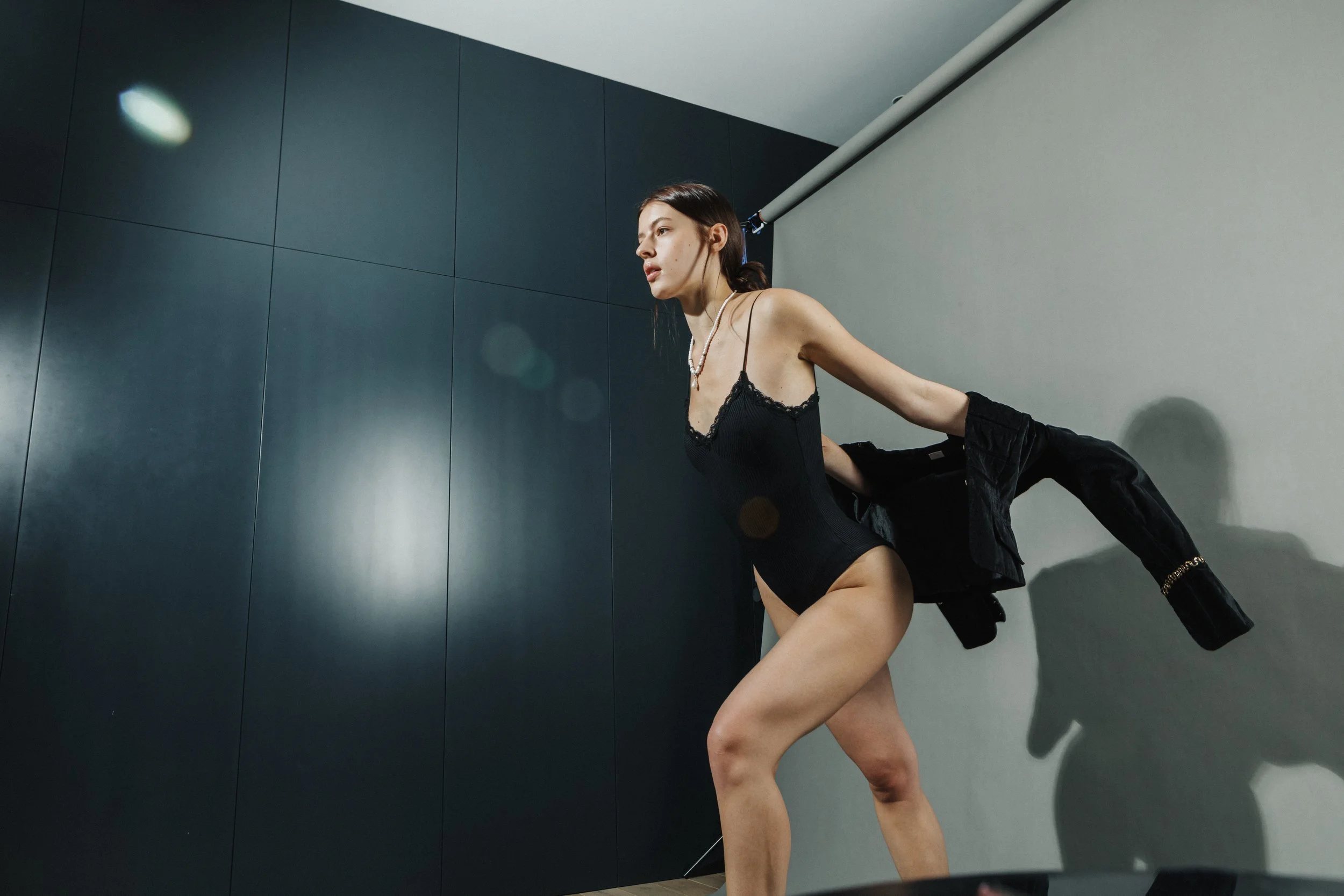 A woman in black lingerie poses against a plain wall, holding a black jacket in one hand, with her shadow cast on the wall.