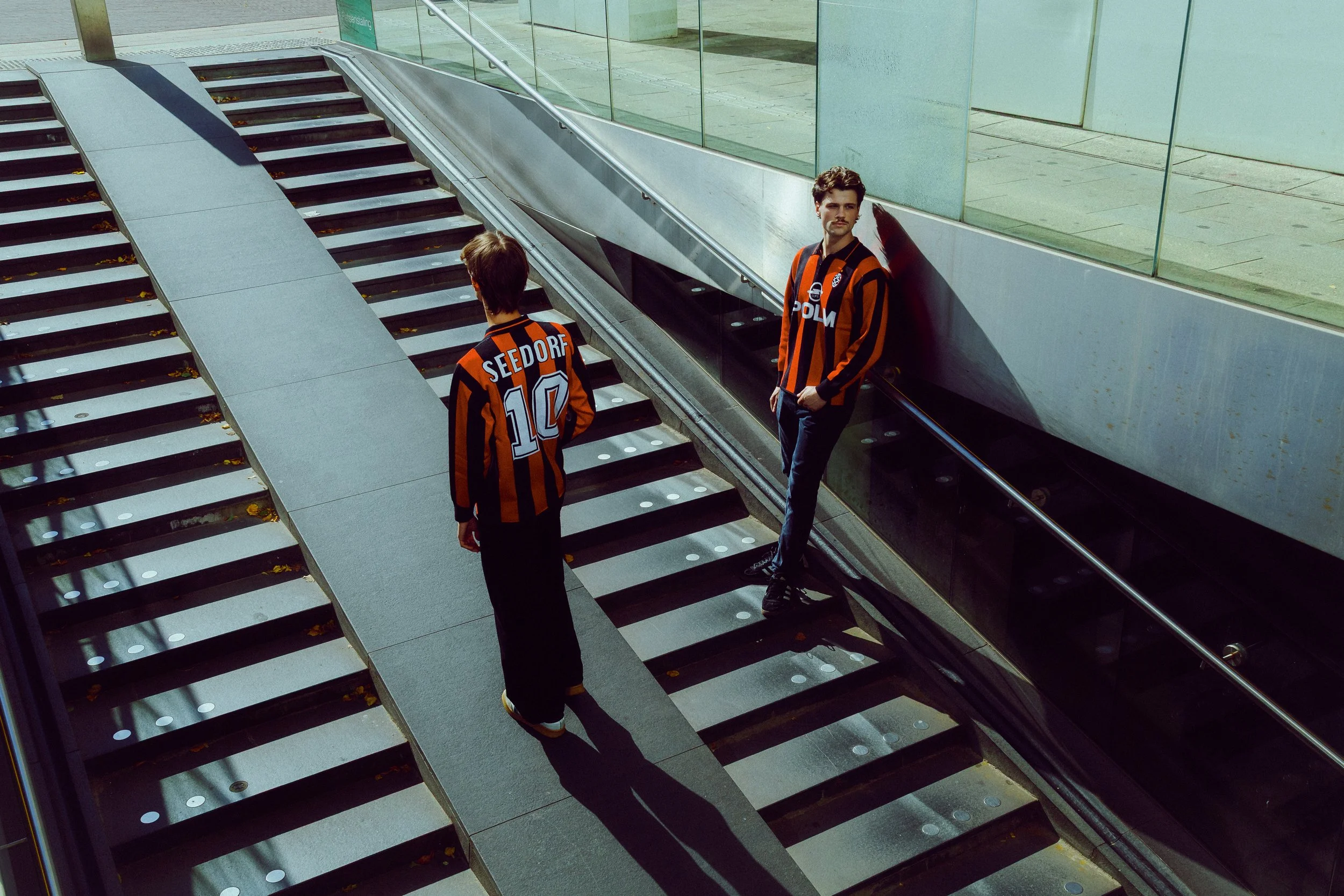 Two men in black and orange striped soccer jerseys are on a modern outdoor staircase, with one walking up the stairs and the other leaning against the wall.