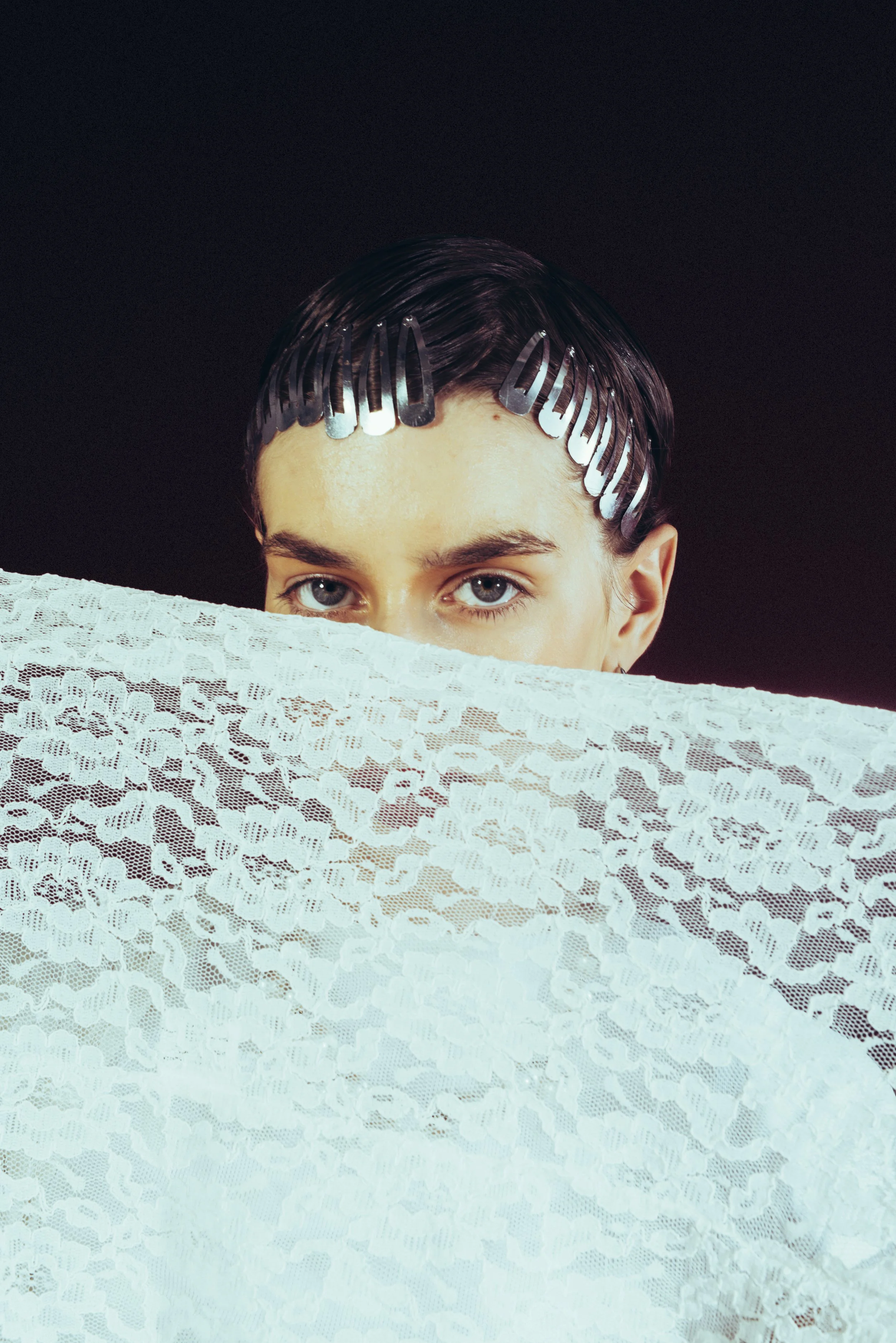 Person with dark hair and blue eyes partially obscured by white lace fabric, holding it up, with hair clips on their head.