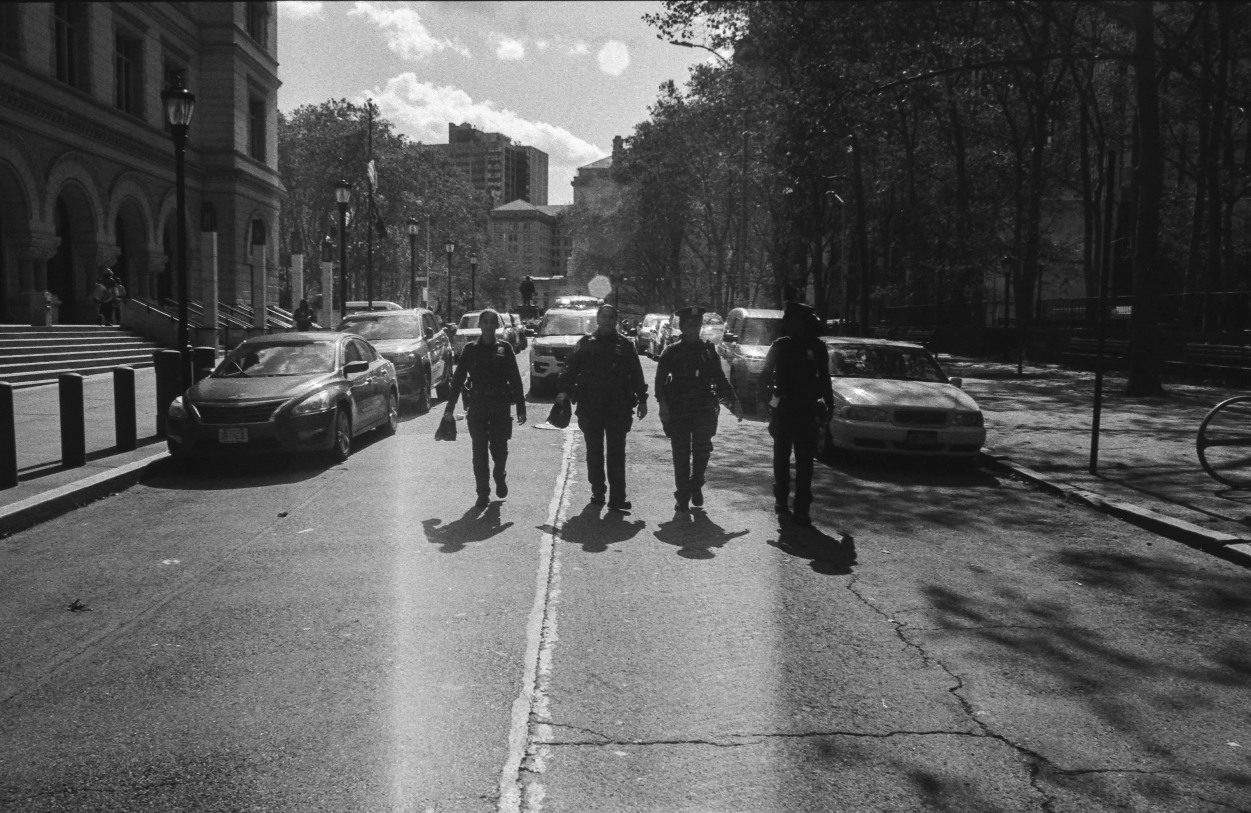Black and white photo of four police officers walking on a city street with parked cars and trees, buildings in the background, on a bright sunny day.